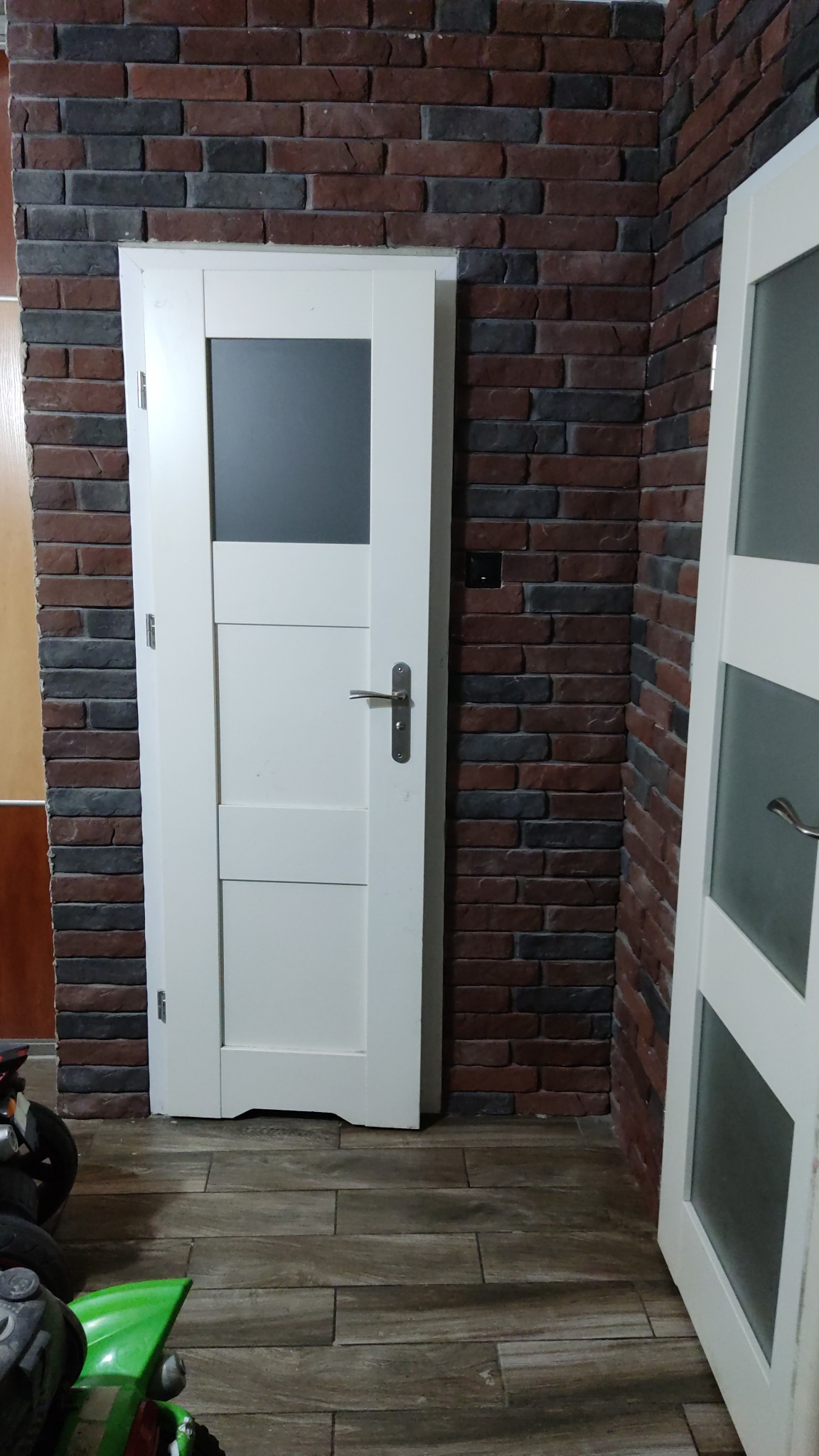 Białe drzwi wewnętrzne z szybą, zamontowane w ścianie z cegły dekoracyjnej, widoczne fragmenty podłogi imitującej drewno oraz częściowo widoczne kolejne drzwi.