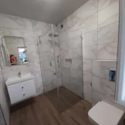 Nowoczesna łazienka z kabiną prysznicową typu walk-in, umywalką z szafką, podwieszaną toaletą i drewnopodobną podłogą. Na ścianach płytki imitujące marmur.