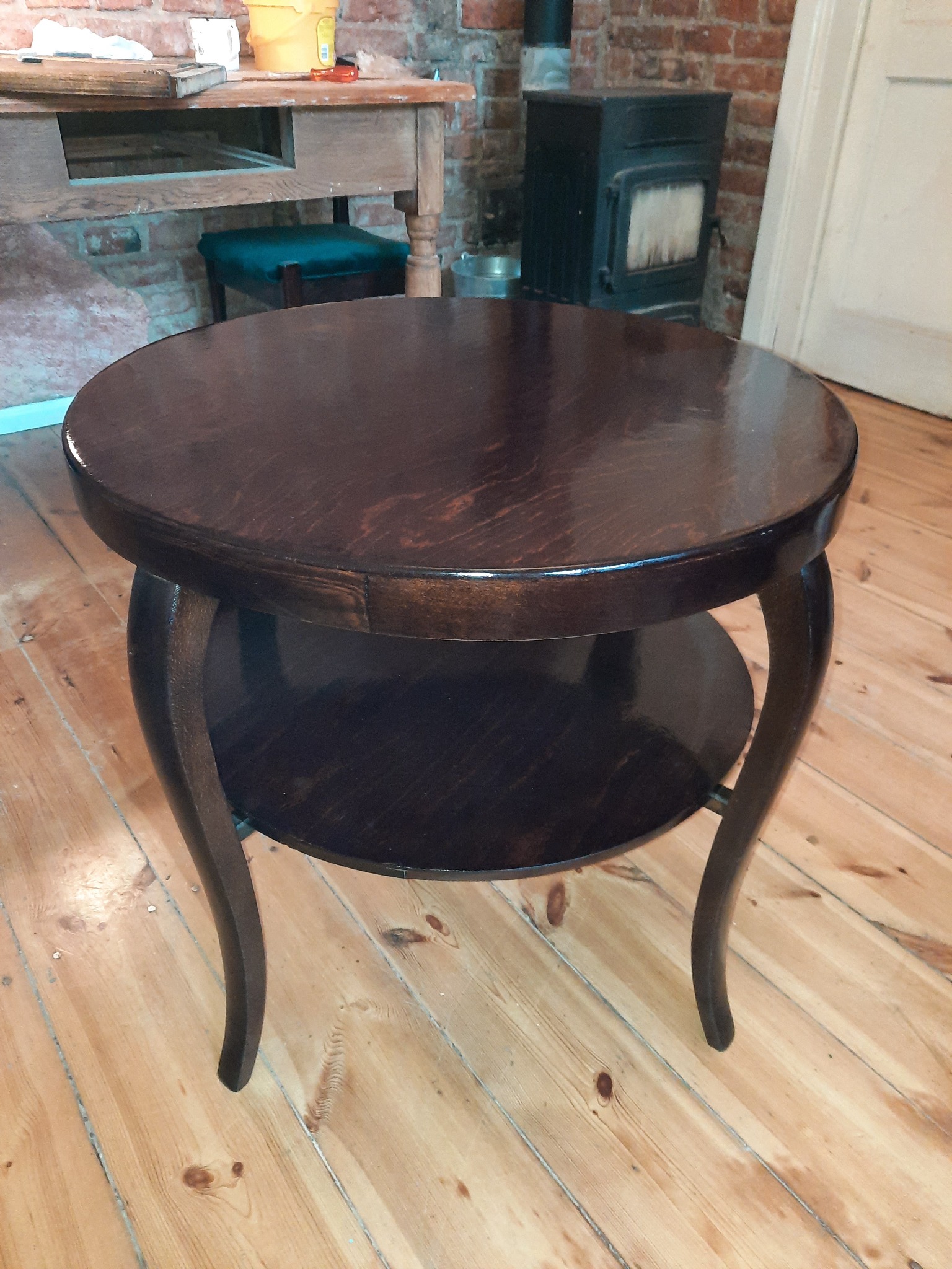 Okrągły, ciemnobrązowy stolik kawowy z półką na giętych nogach, stojący na drewnianej podłodze, z piecem kaflowym i ceglaną ścianą w tle.