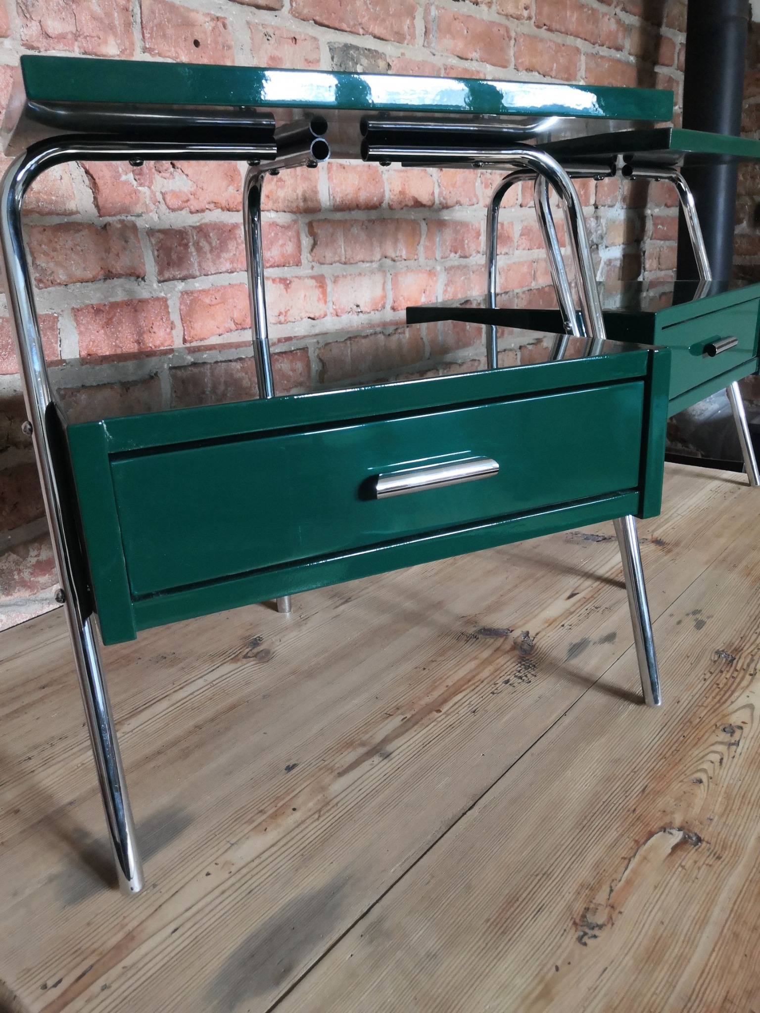 Odnowiony, lakierowany na butelkową zieleń stolik nocny z chromowanymi nogami i uchwytami szuflad, na tle ściany z czerwonej cegły i drewnianej podłogi.