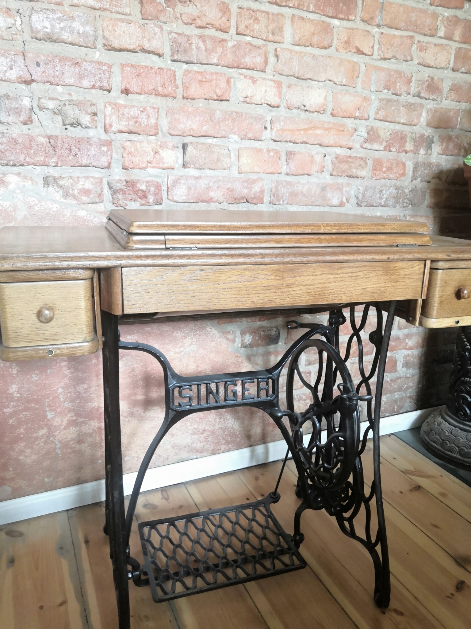 Antyczna maszyna do szycia Singer z drewnianym blatem i szufladami, ustawiona na tle ceglanej ściany, z widocznym żeliwnym mechanizmem i pedałem.