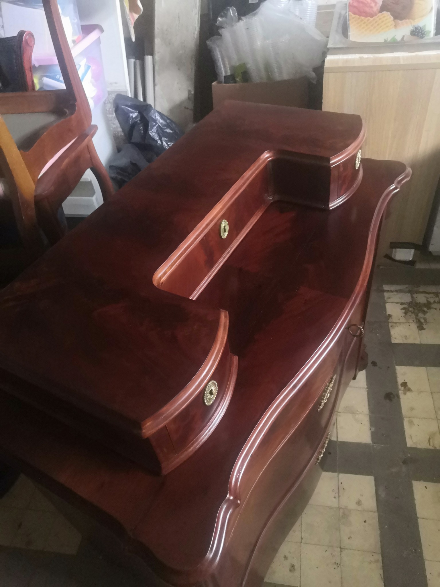 Odrestaurowana, lakierowana na wysoki połysk, mahoniowa toaletka z ozdobnymi, mosiężnymi uchwytami i zamkami, ustawiona w pomieszczeniu z widoczną podłogą w kratkę i elementami wyposażenia w tle.