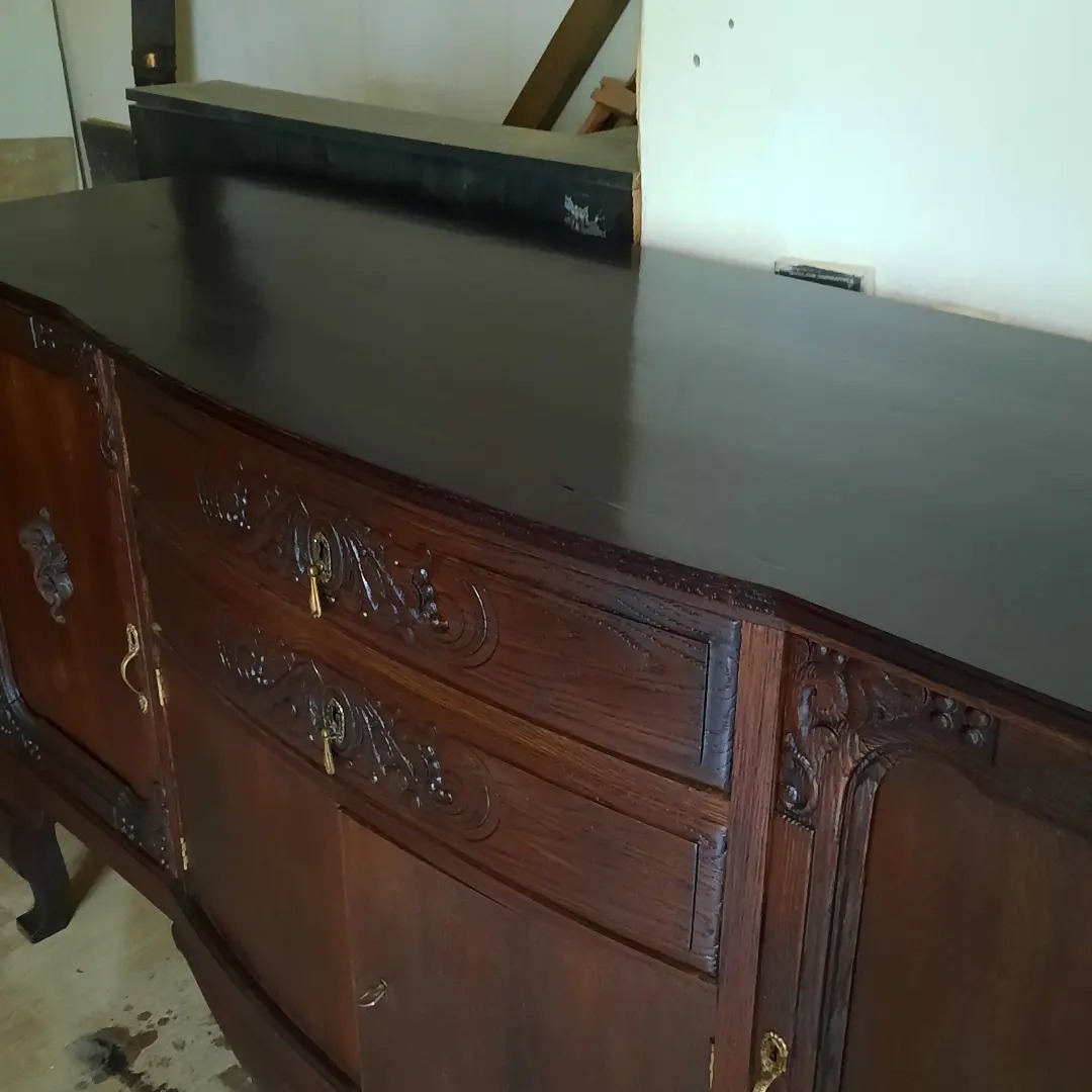 Antyczny, ciemnobrązowy kredens z rzeźbionymi detalami, po renowacji, z widocznymi śladami pracy na podłodze.