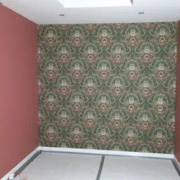 Pokój po odświeżeniu: jedna ściana pomalowana na matowy róż, druga wytapetowana w zielono-różowe, symetryczne wzory roślinne. Podłoga zabezpieczona białym kartonem, widoczna lampa budowlana przy...