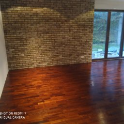 Marwit Vitalii Somushkin - Jasnobrązowa ściana z cegły dekoracyjnej, parkiet z ciemnego drewna w pustym pokoju z dużymi oknami tarasowymi wychodzącymi na zieleń. Na zewnątrz widoczne materiały budowlane zabezpieczone folią.