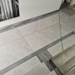 Marwit Vitalii Somushkin - Jasnoszare płytki podłogowe ułożone w kwadraty z ciemniejszymi szarymi obramowaniami, widoczne rury instalacyjne w ścianie i fragment schodów z wykończeniem z płytek.