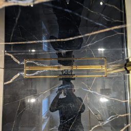 Marwit Vitalii Somushkin - Czarne, połyskujące płytki z białymi i złotymi żyłkami, zamontowane w kabinie prysznicowej z odpływem liniowym w złotym kolorze; widoczne odbicie osoby robiącej zdjęcie.