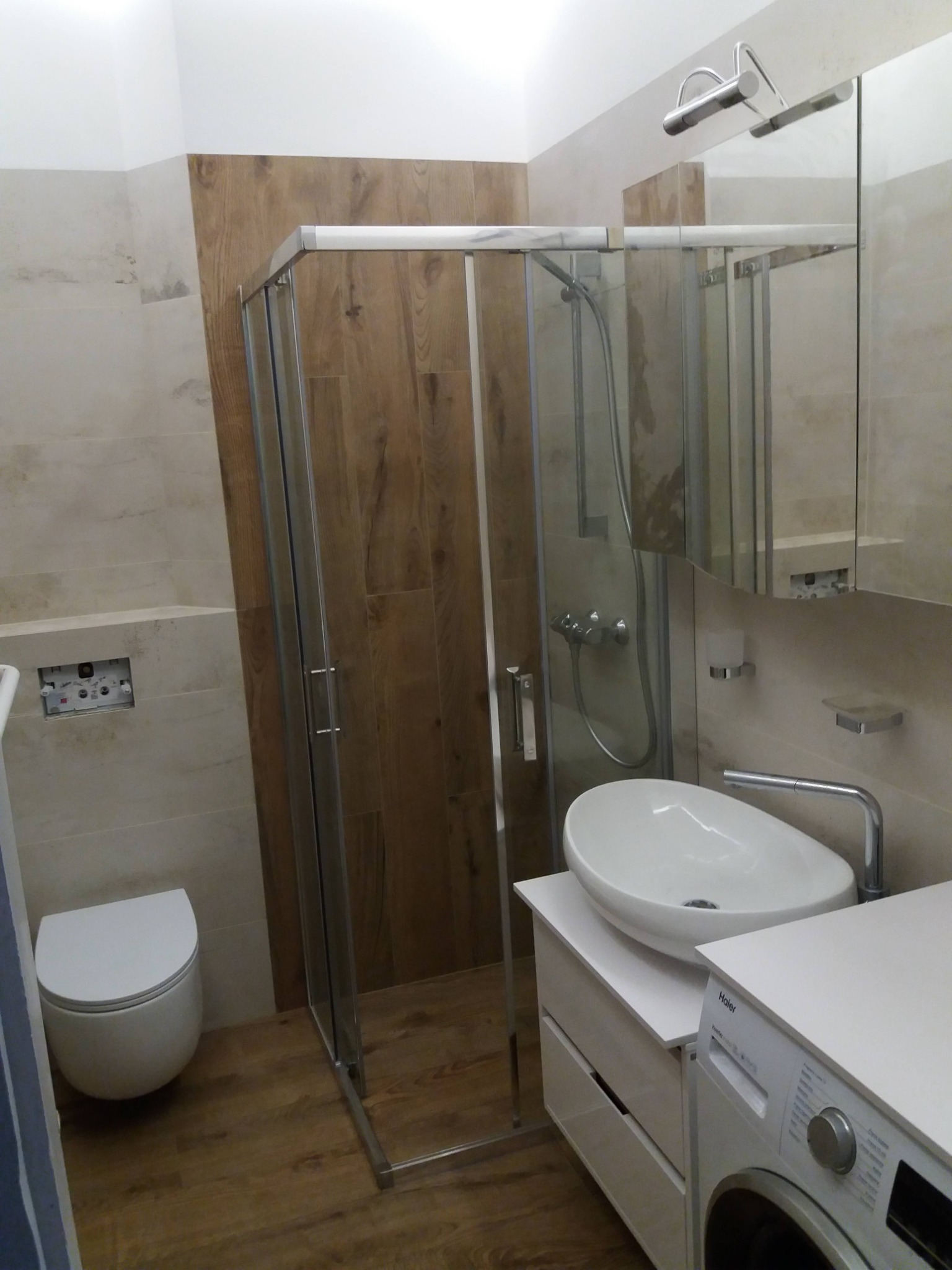 Wykończona łazienka z kabiną prysznicową, umywalką nablatową i pralką, połączenie płytek imitujących drewno i beżowych płytek ceramicznych.