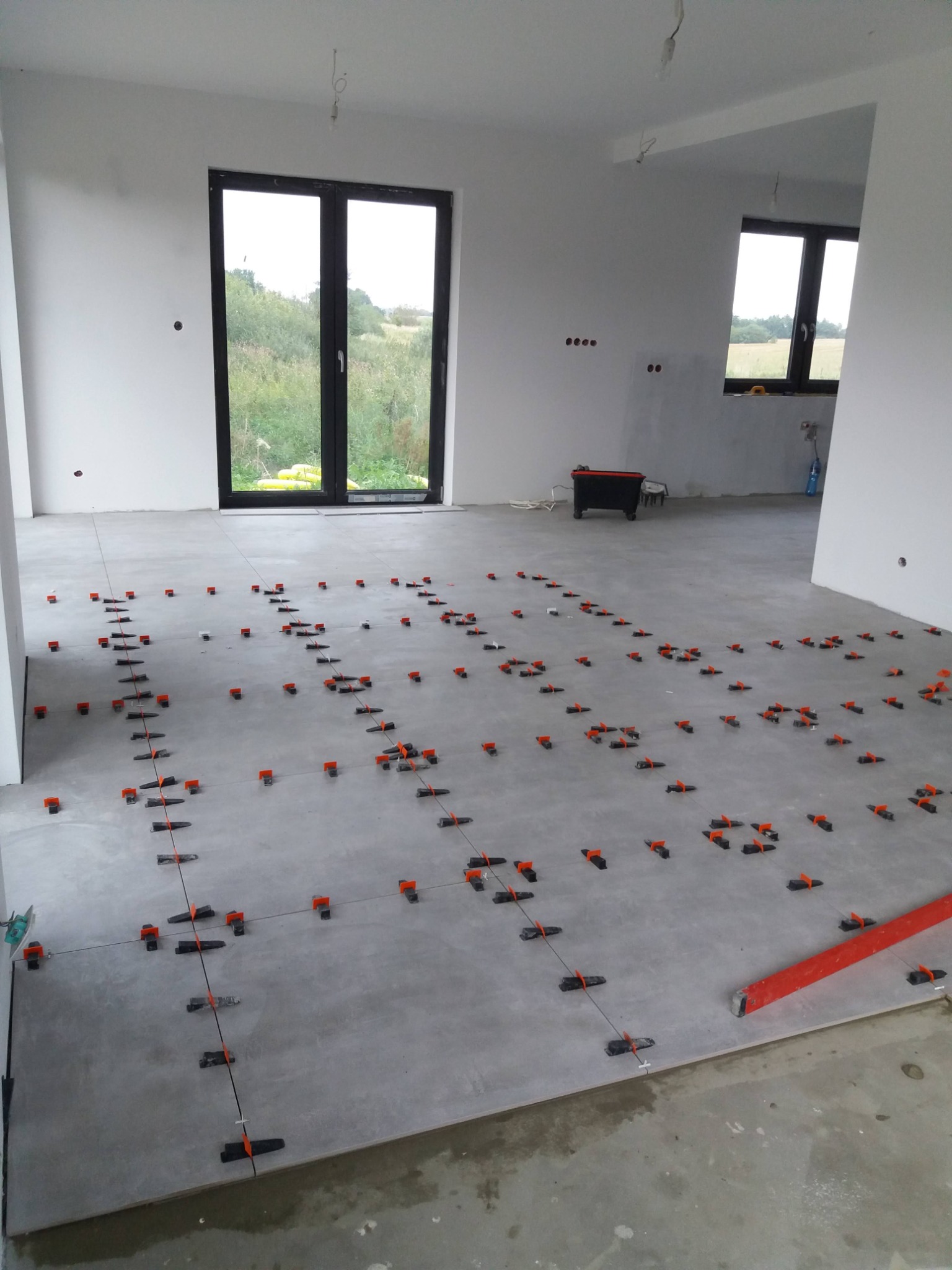 Układanie szarych płytek podłogowych z użyciem systemu poziomowania w nowym, nieumeblowanym pomieszczeniu z dużymi oknami.