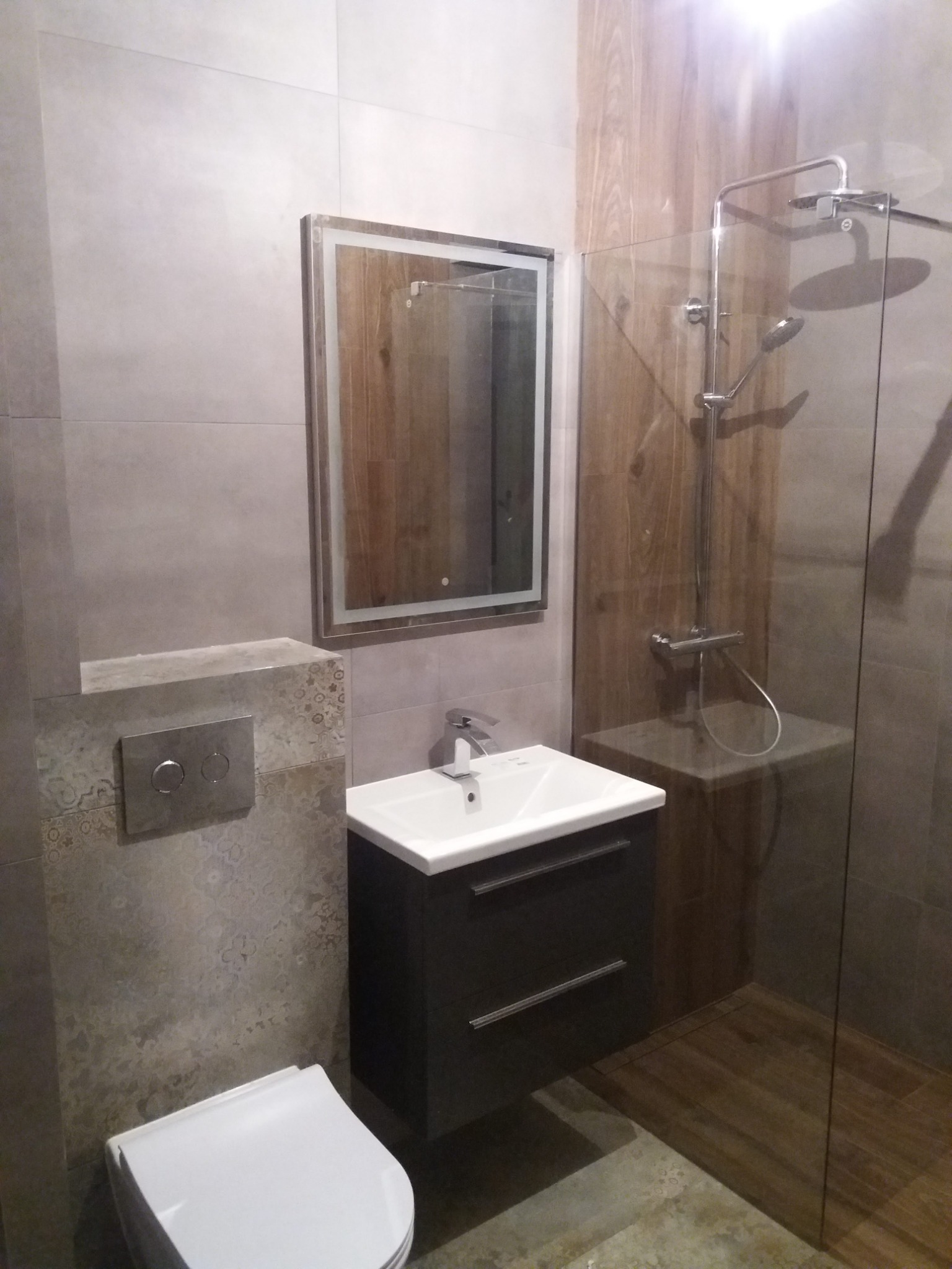 Nowoczesna łazienka z kabiną prysznicową typu walk-in, umywalką z szafką w kolorze grafitowym, podświetlanym lustrem i płytkami imitującymi drewno na ścianie w strefie prysznica.