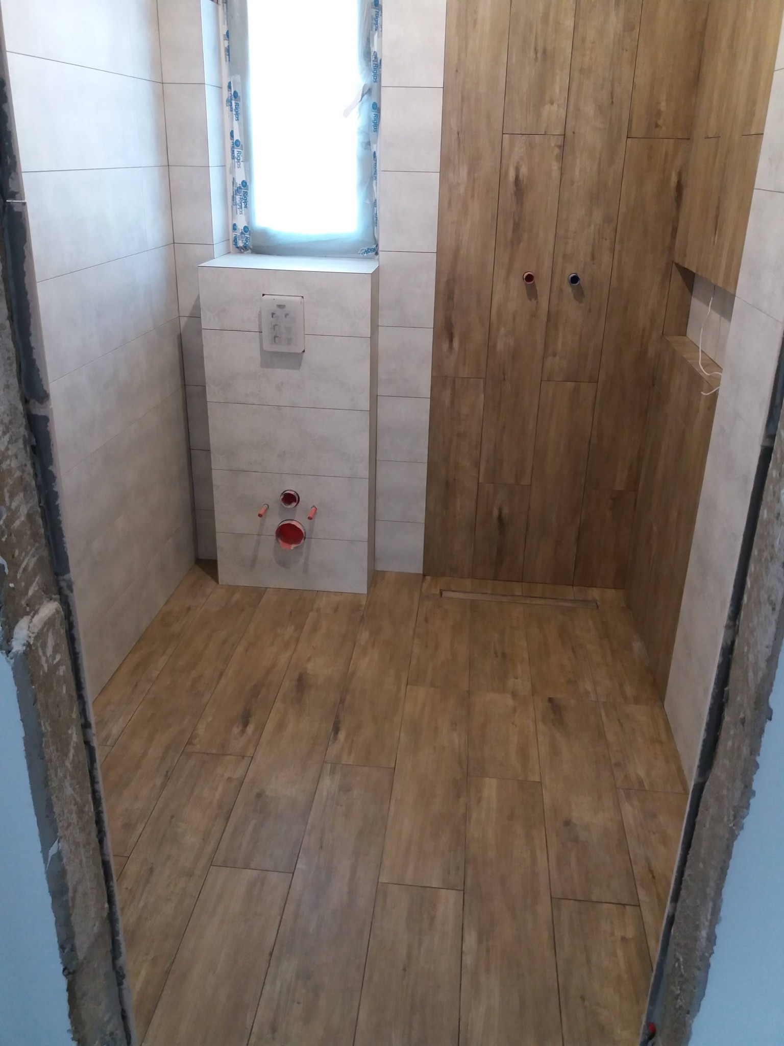 Wnętrze łazienki w trakcie wykańczania, z drewnopodobnymi płytkami na podłodze i ścianie prysznicowej, widoczne przyłącza wodne i fragment okna z folią ochronną.