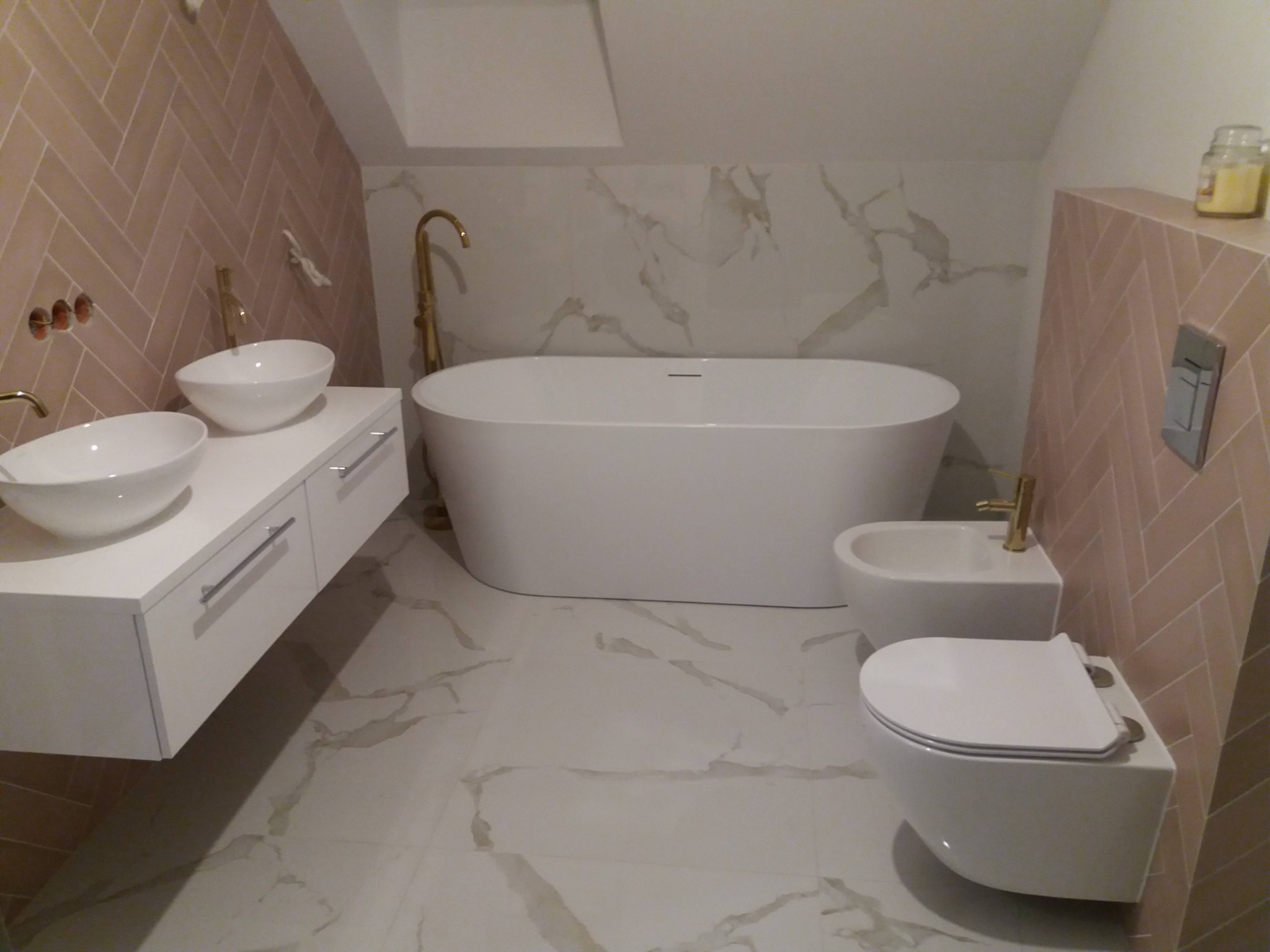 Nowoczesna łazienka na poddaszu z wanną wolnostojącą, umywalkami nablatowymi na białej szafce, bidetem i toaletą podwieszaną, wykończona płytkami w odcieniach różu i bieli z marmurkowym wzorem...