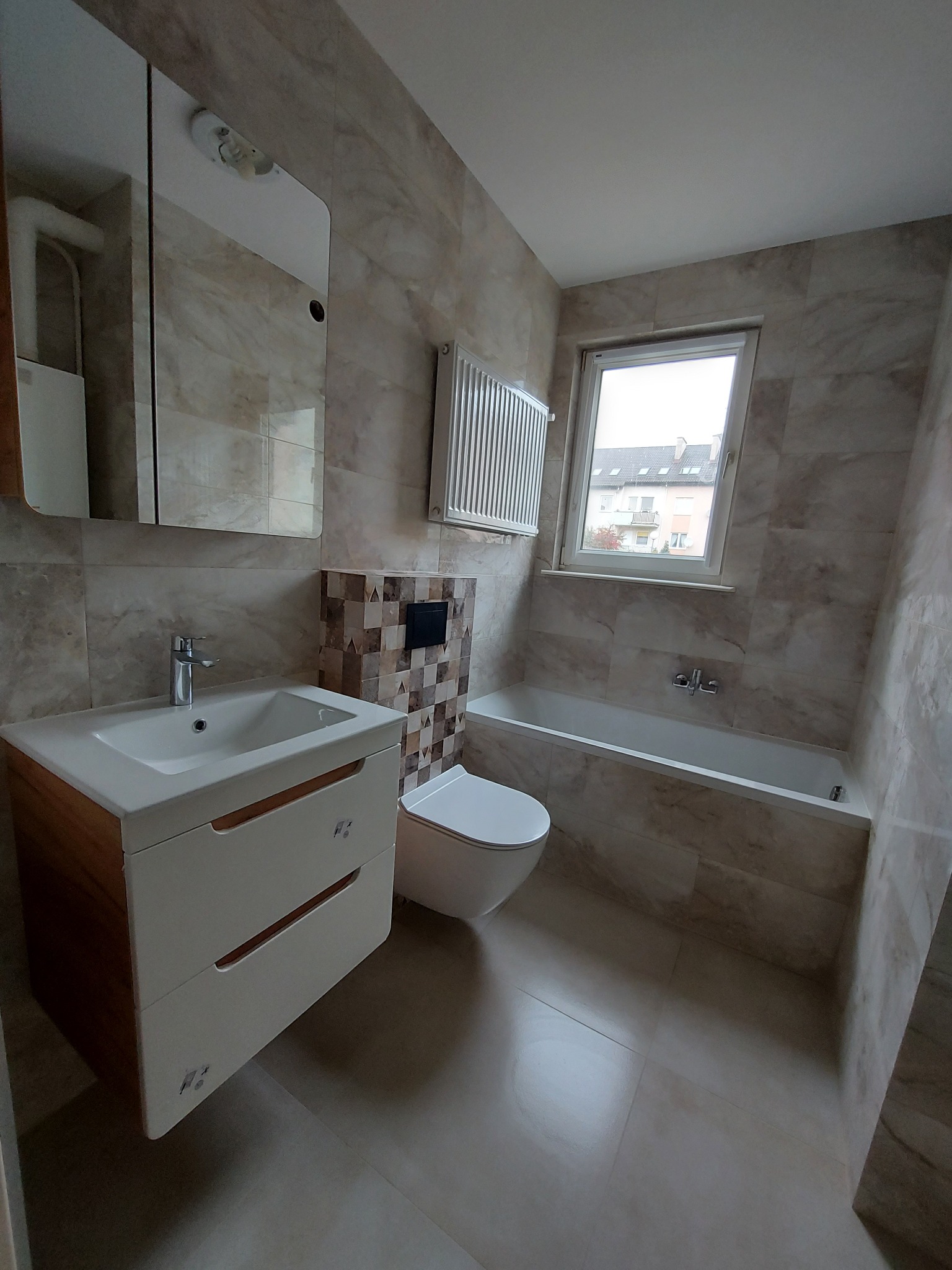 Jasna łazienka z beżowymi płytkami imitującymi marmur, wyposażona w umywalkę z szafką, toaletę z zabudową oraz wannę pod oknem.