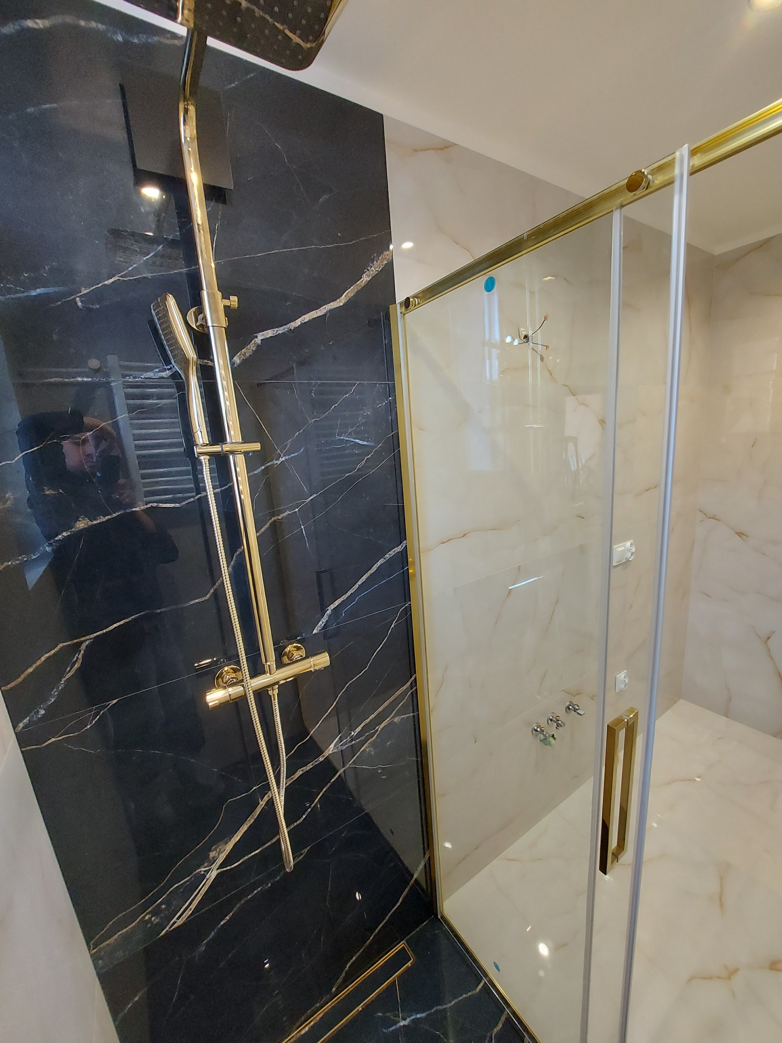 Luksusowa kabina prysznicowa z czarnymi marmurowymi ścianami i złotą armaturą, przesuwne drzwi kabiny z złotymi ramami, jasne marmurowe płytki na podłodze i ścianach wewnątrz kabiny.