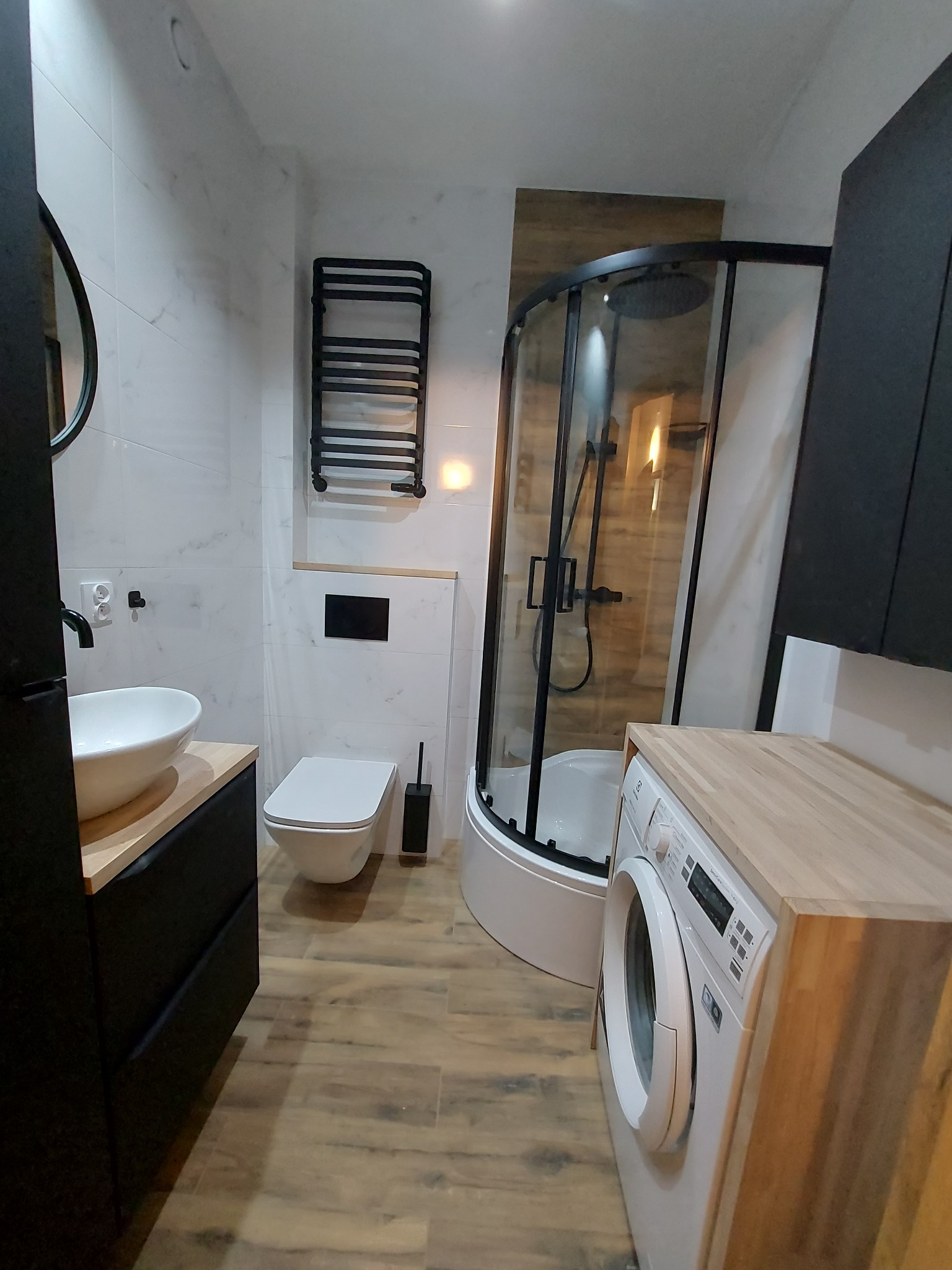 Nowoczesna łazienka z kabiną prysznicową typu walk-in, wiszącą toaletą, umywalką nablatową na czarnej szafce, grzejnikiem drabinkowym i pralką zabudowaną blatem z jasnego drewna.