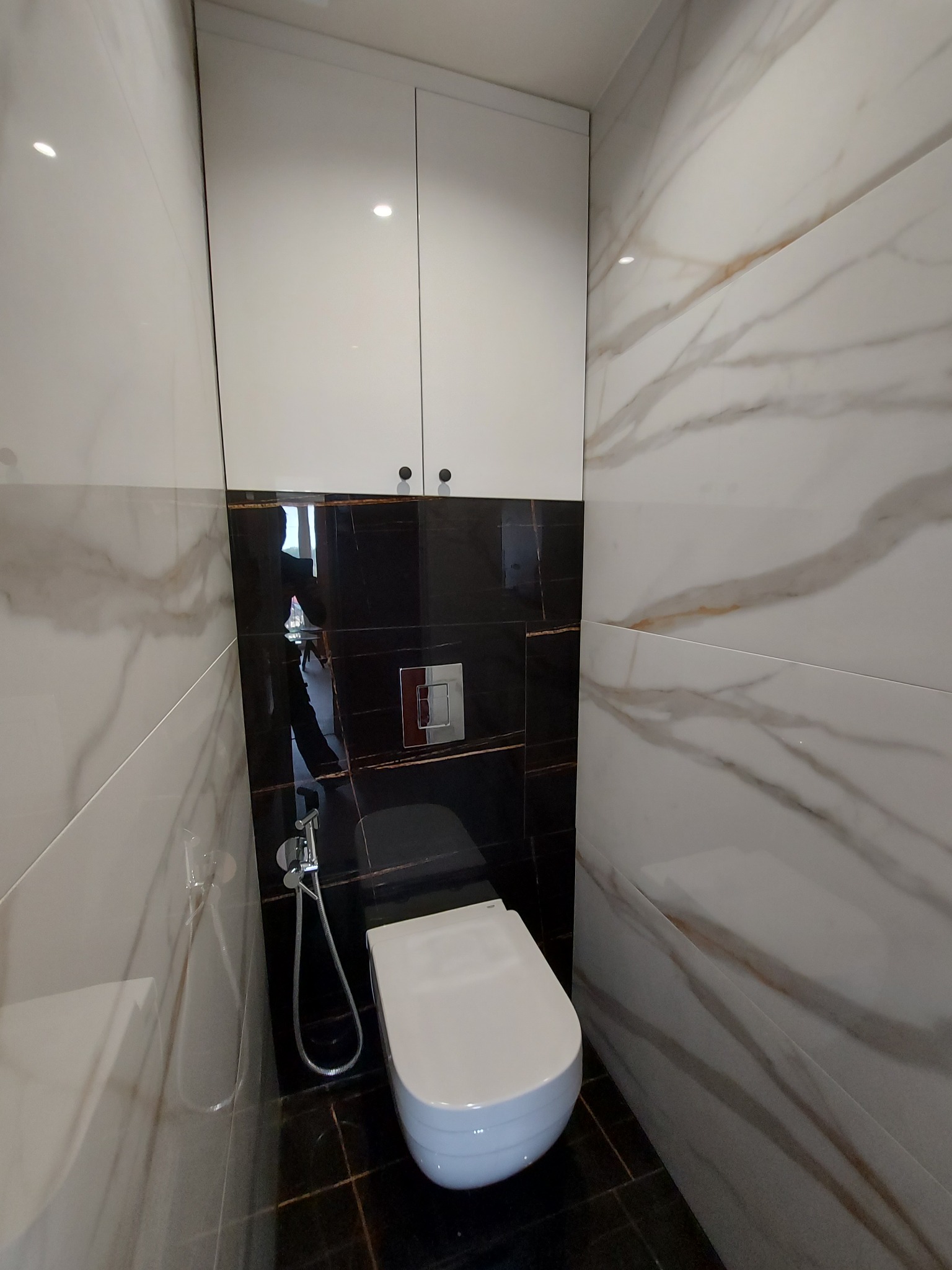 Nowoczesna toaleta podwieszana z bidetką, obudowana czarnymi i białymi płytkami imitującymi marmur, z szafką wiszącą nad nią.