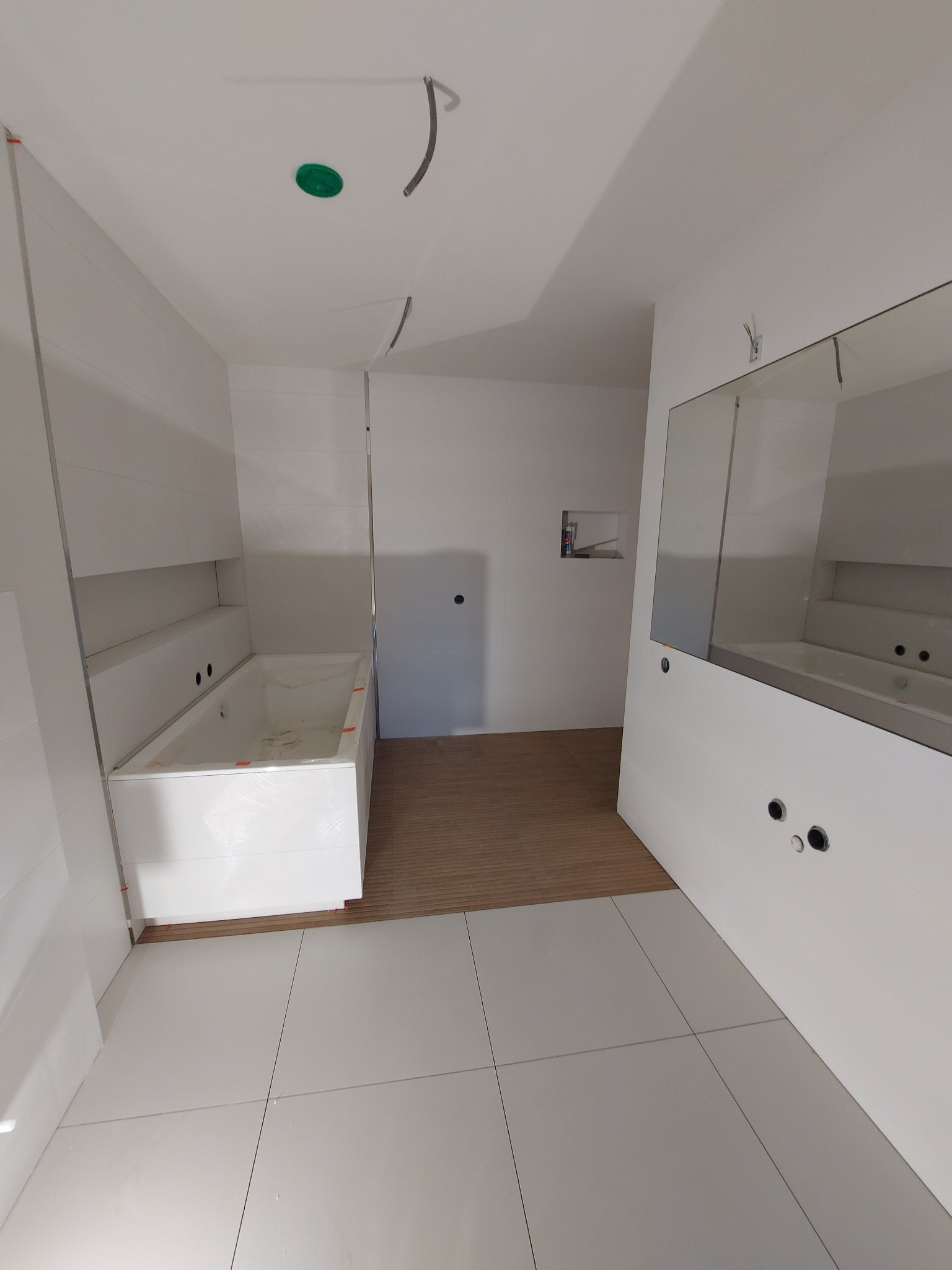 Wnętrze łazienki w trakcie wykańczania: białe płytki na ścianach i podłodze, zabudowana wanna w folii ochronnej, widoczne instalacje elektryczne i wodne, drewnopodobna podłoga w przejściu.