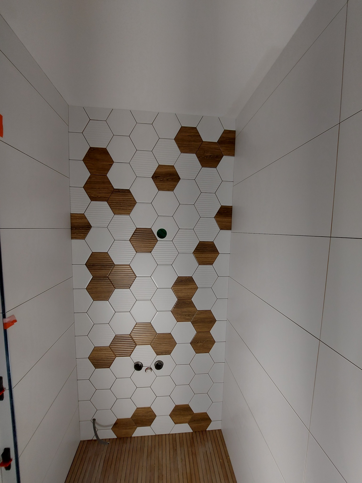 Ściana w kabinie prysznicowej wyłożona białymi i drewnopodobnymi heksagonalnymi płytkami, z drewnianą podłogą i widocznymi otworami na armaturę.