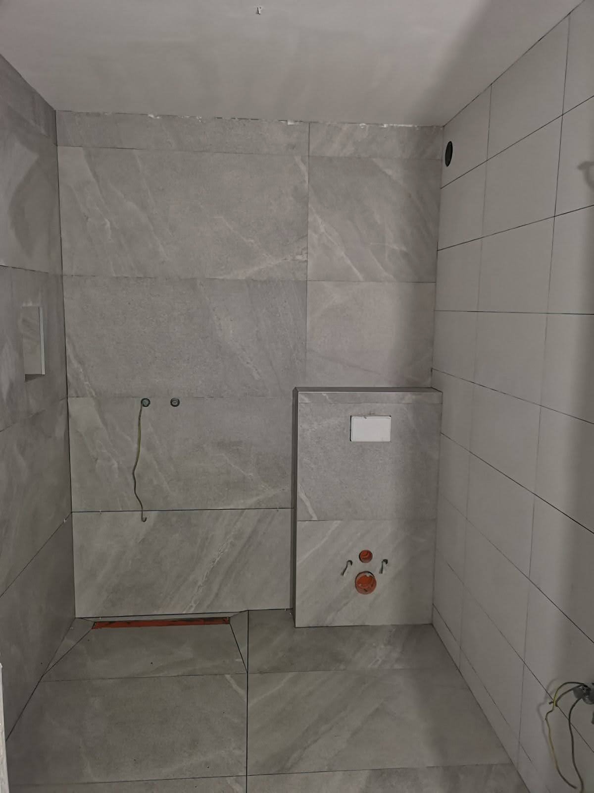 Wnętrze łazienki w trakcie prac wykończeniowych, widoczne szare płytki imitujące kamień na ścianach oraz podłodze, zabudowa stelaża podtynkowego WC z widocznymi przyłączami hydraulicznymi...