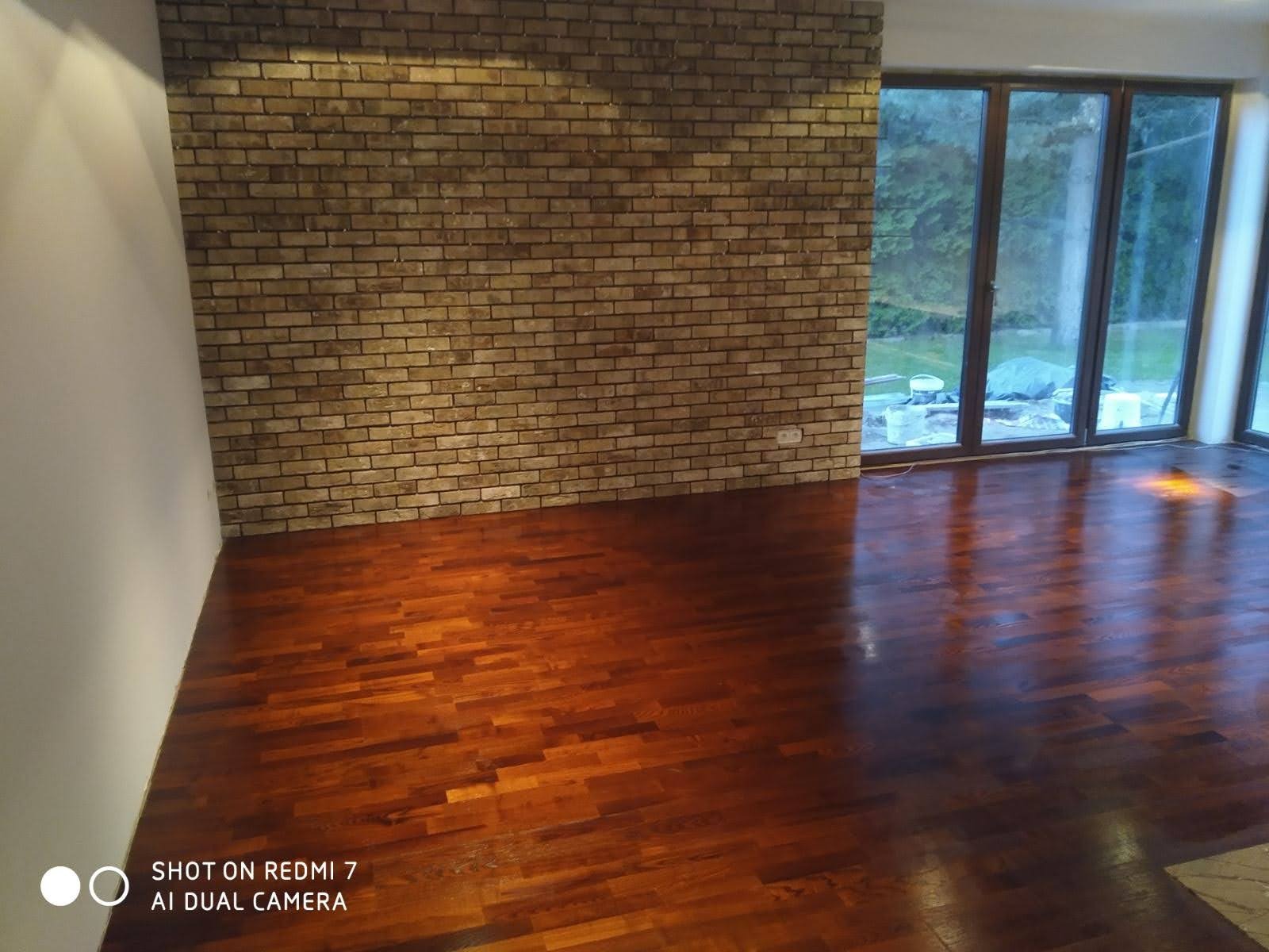 Jasnobrązowa ściana z cegły dekoracyjnej, parkiet z ciemnego drewna w pustym pokoju z dużymi oknami tarasowymi wychodzącymi na zieleń. Na zewnątrz widoczne materiały budowlane zabezpieczone folią.