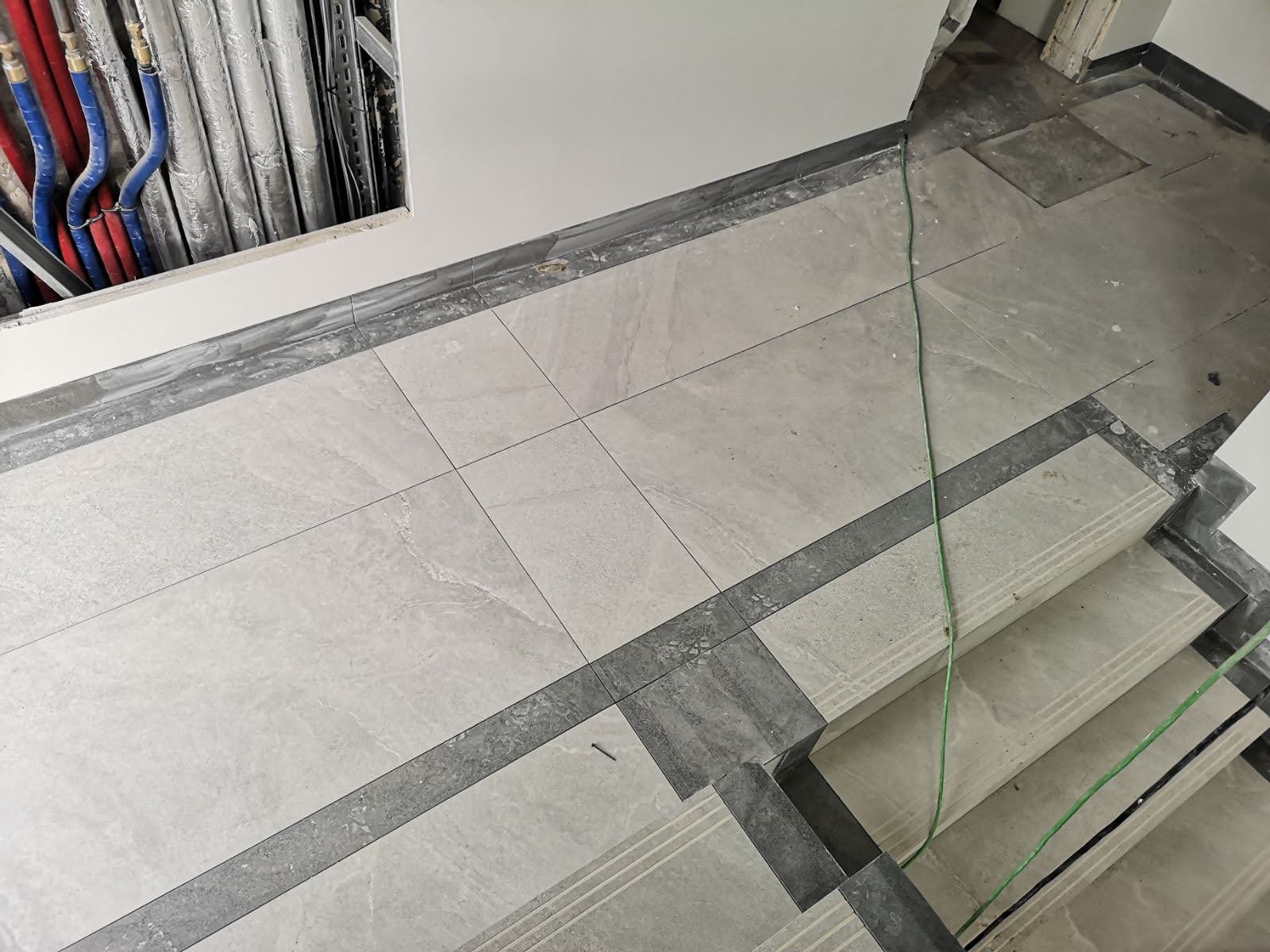 Jasnoszare płytki podłogowe ułożone w kwadraty z ciemniejszymi szarymi obramowaniami, widoczne rury instalacyjne w ścianie i fragment schodów z wykończeniem z płytek.