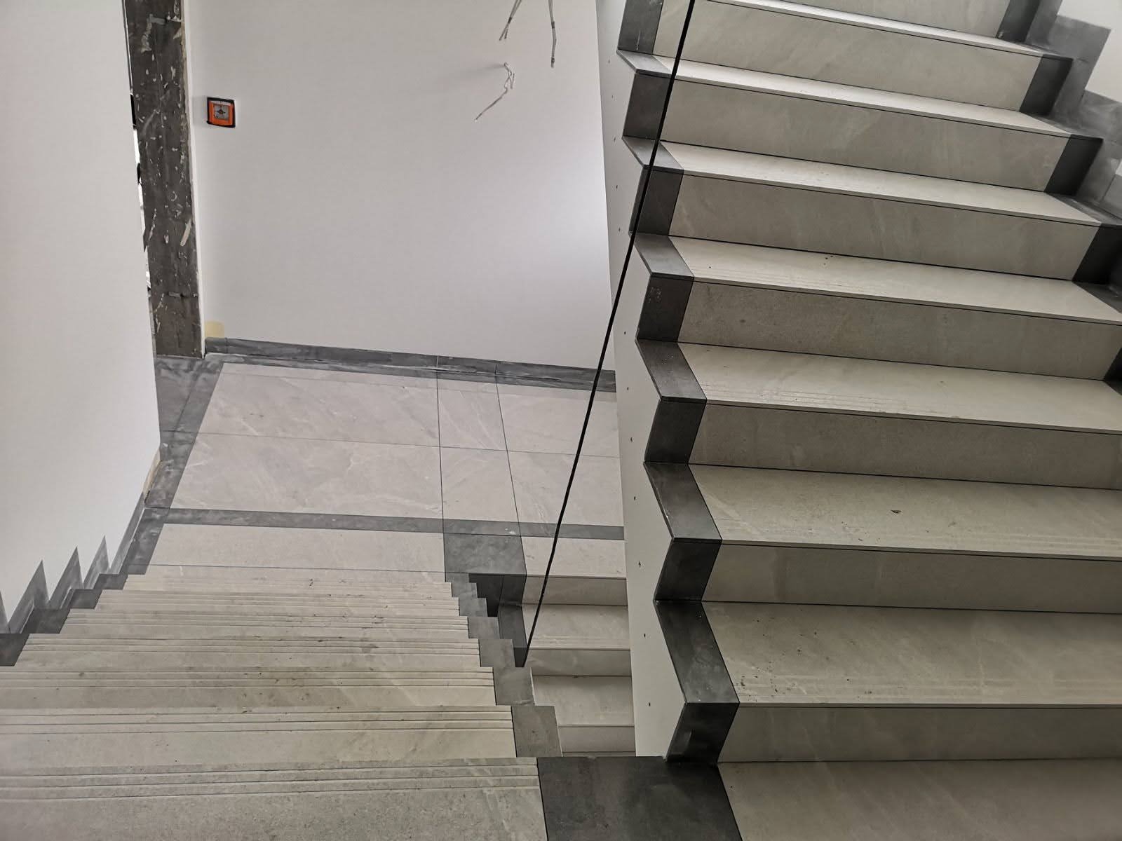 Wykończone schody z jasnych płytek z ciemnymi krawędziami, widok z góry, szklana balustrada, na podłodze płytki w tym samym stylu.