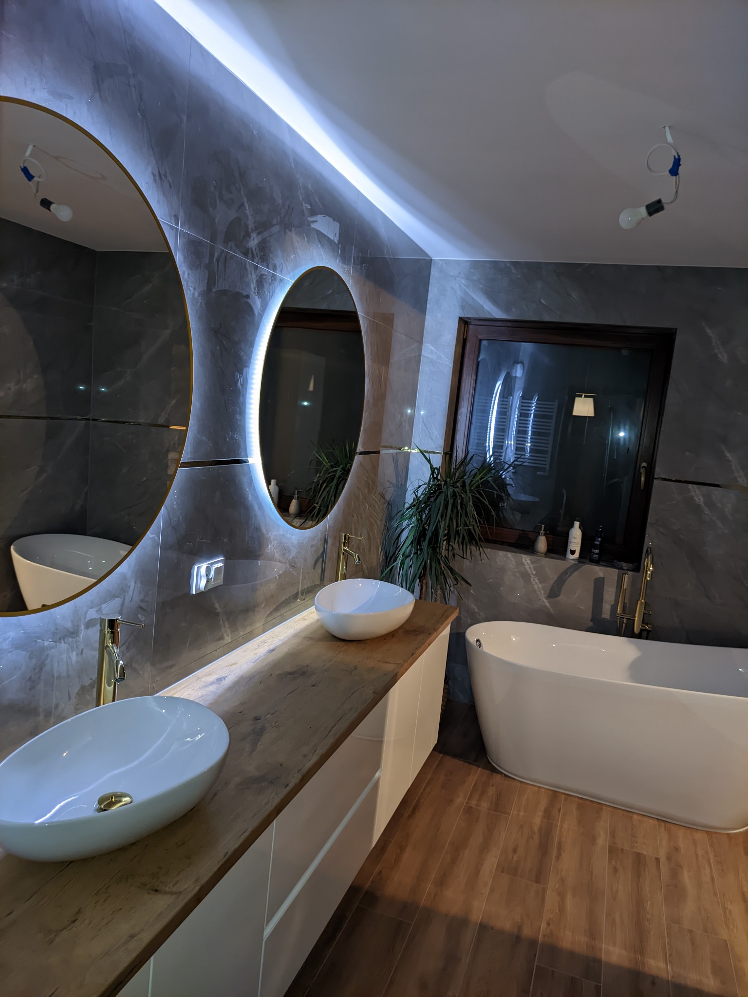 Nowoczesna łazienka z dwoma okrągłymi lustrami podświetlanymi LED, drewnianym blatem pod umywalki nablatowe, wolnostojącą wanną i szarymi płytkami imitującymi marmur na ścianach.