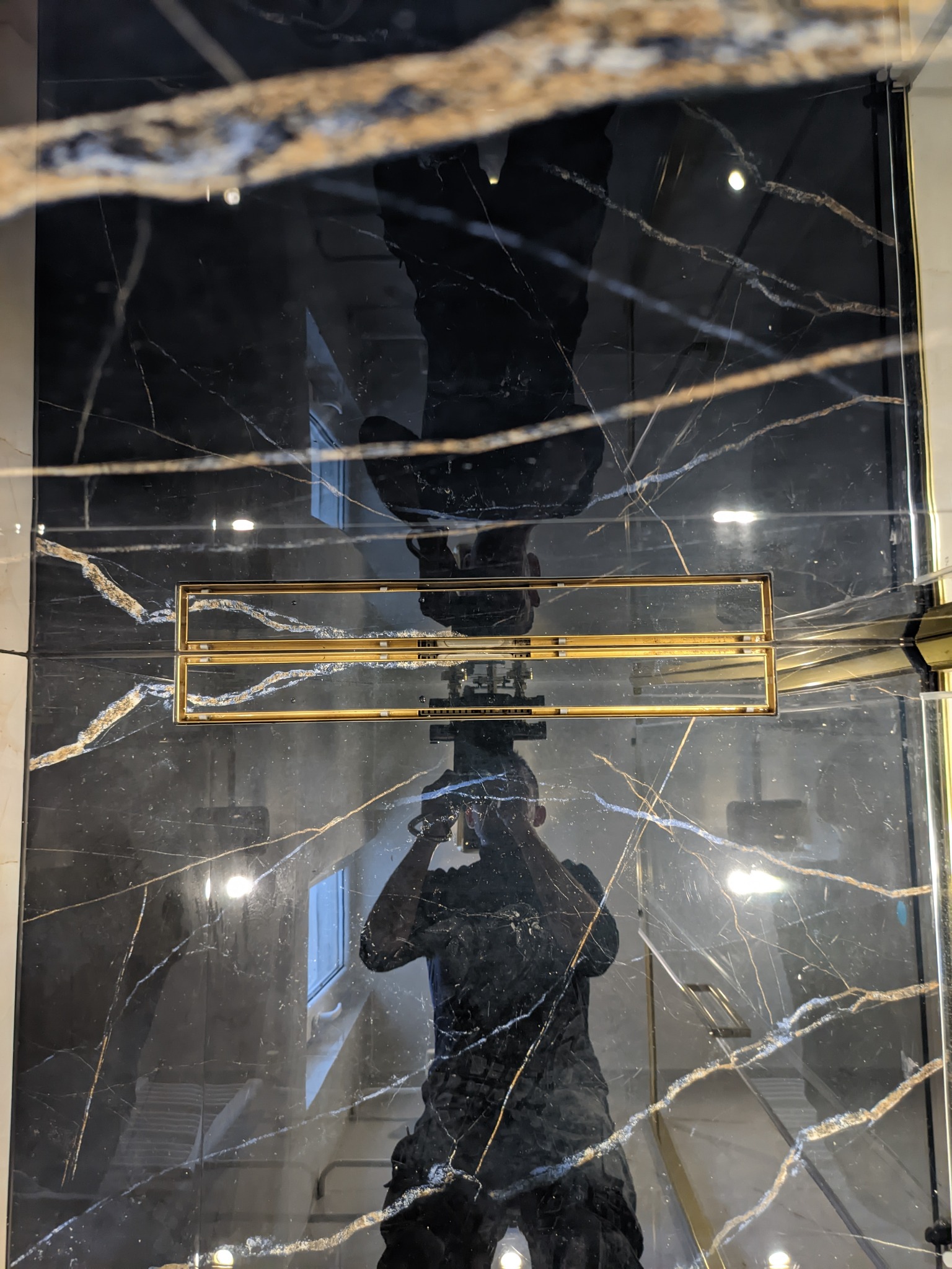 Czarne, połyskujące płytki z białymi i złotymi żyłkami, zamontowane w kabinie prysznicowej z odpływem liniowym w złotym kolorze; widoczne odbicie osoby robiącej zdjęcie.