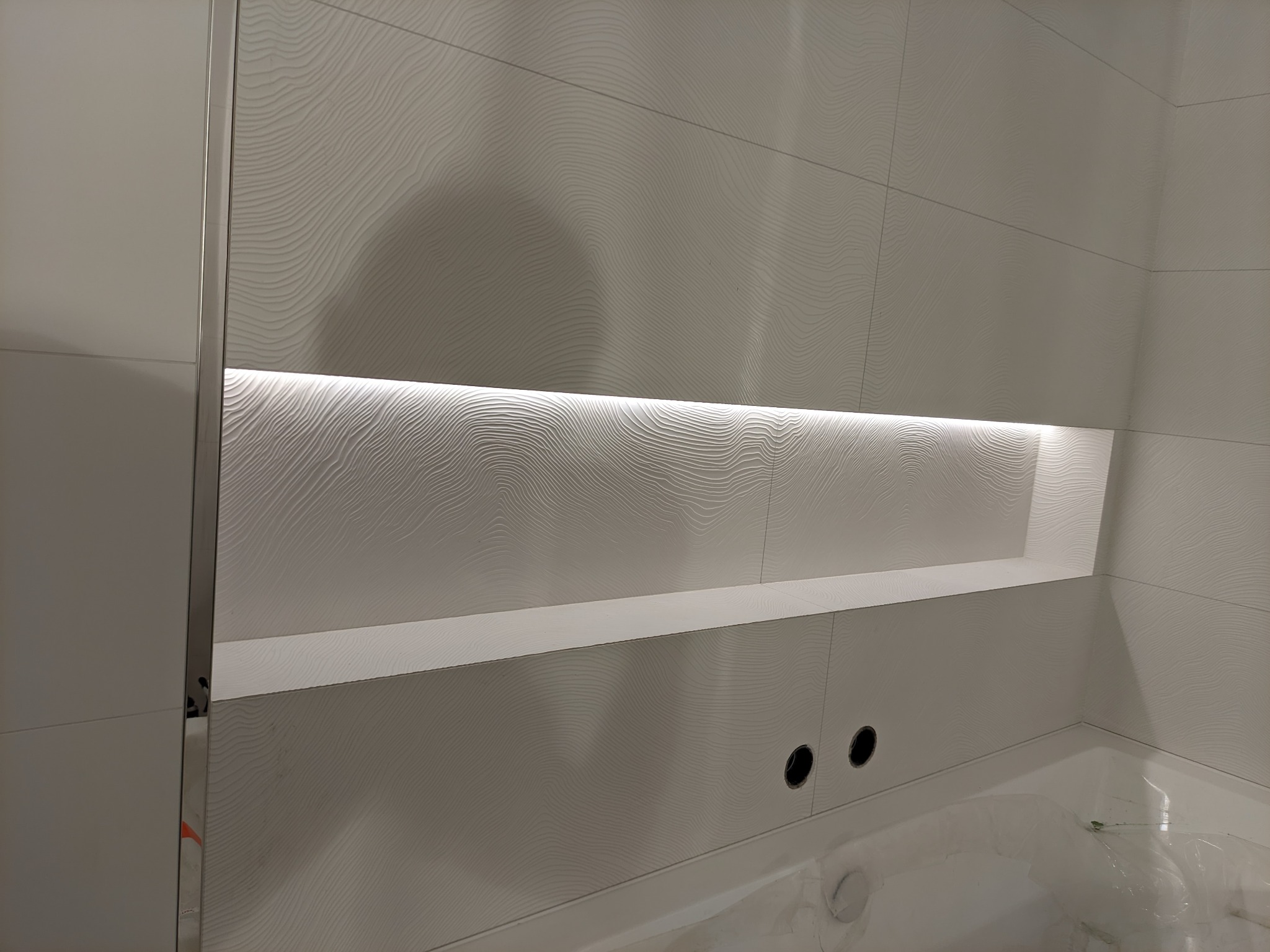 Wykończona ściana w łazience z białymi, falistymi płytkami, podświetlona wnęka i fragment wanny.
