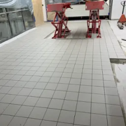 Układanie jasnoszarych, kwadratowych płytek podłogowych w garażu, z dwiema czerwonymi podnośnikami samochodowymi w tle, zabezpieczone folią ściany i brama garażowa.