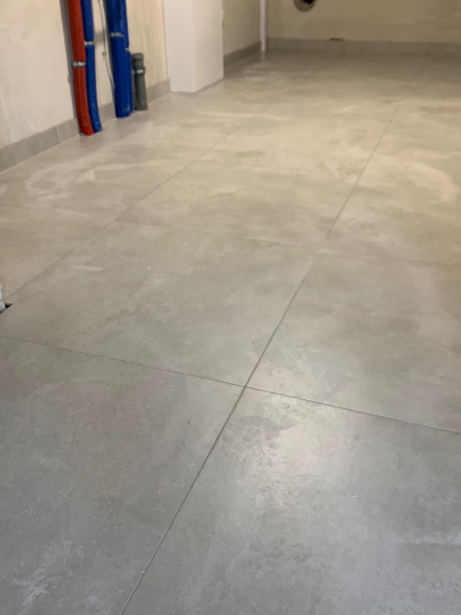 Szare, matowe płytki podłogowe ułożone w dużej przestrzeni, widoczne rury instalacyjne przy ścianie, surowy stan wykończenia.