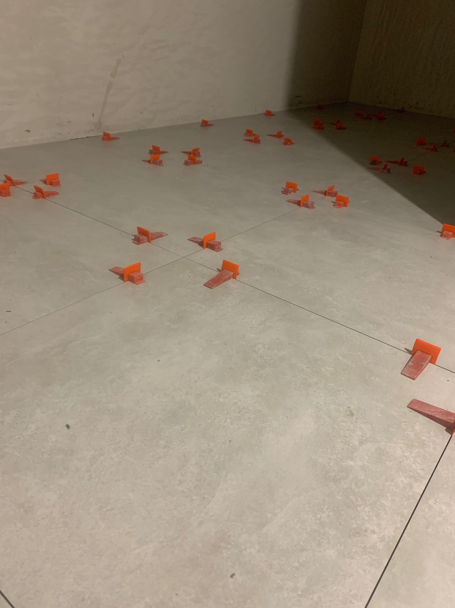 Szare płytki podłogowe z pomarańczowymi klipsami poziomującymi, widoczne czarne linie fug, w tle ściana w surowym stanie.