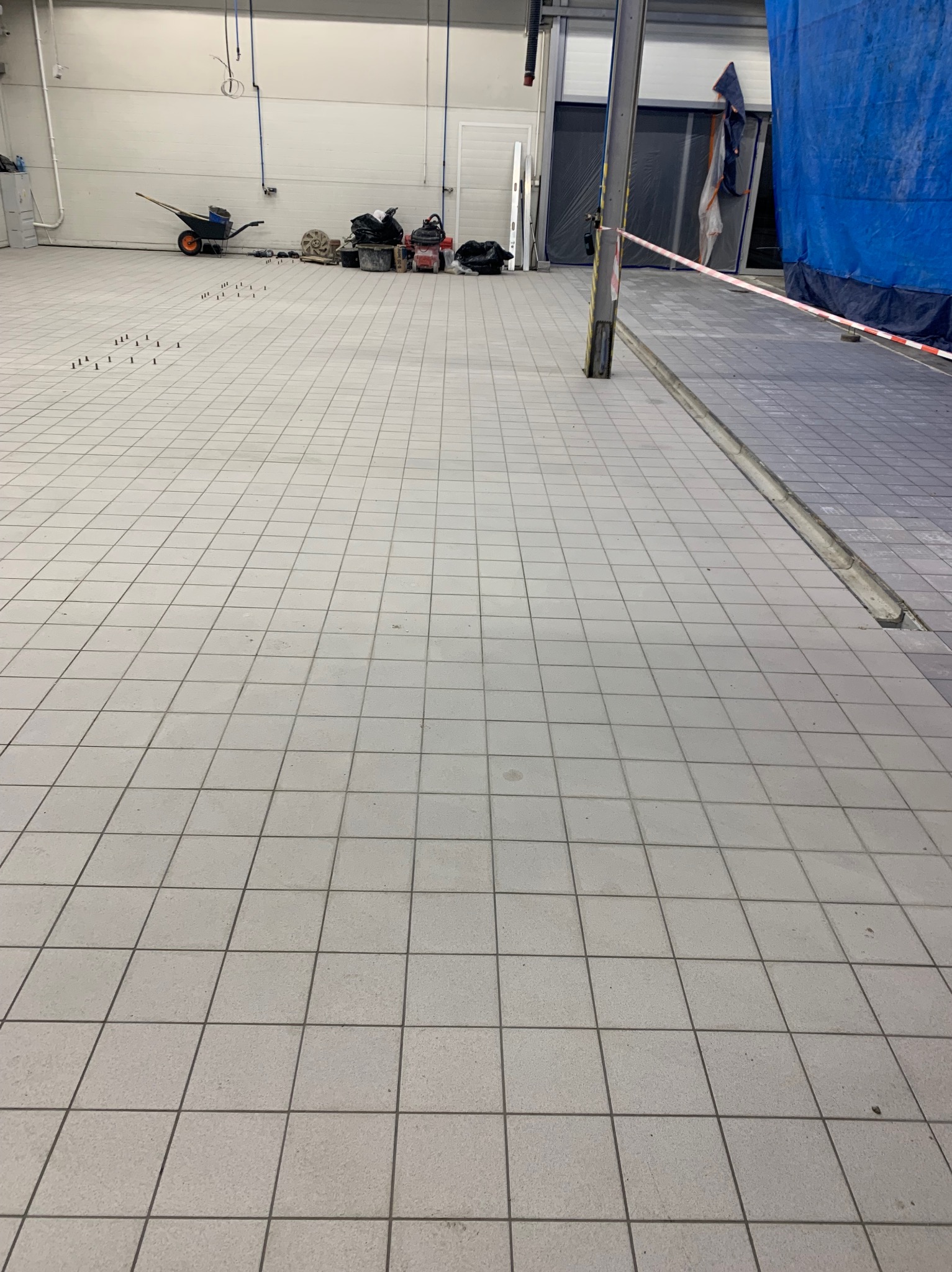 Jasnoszare płytki podłogowe w dużym pomieszczeniu, widoczne narzędzia i materiały budowlane w tle, małe kołki w rzędach na podłodze.