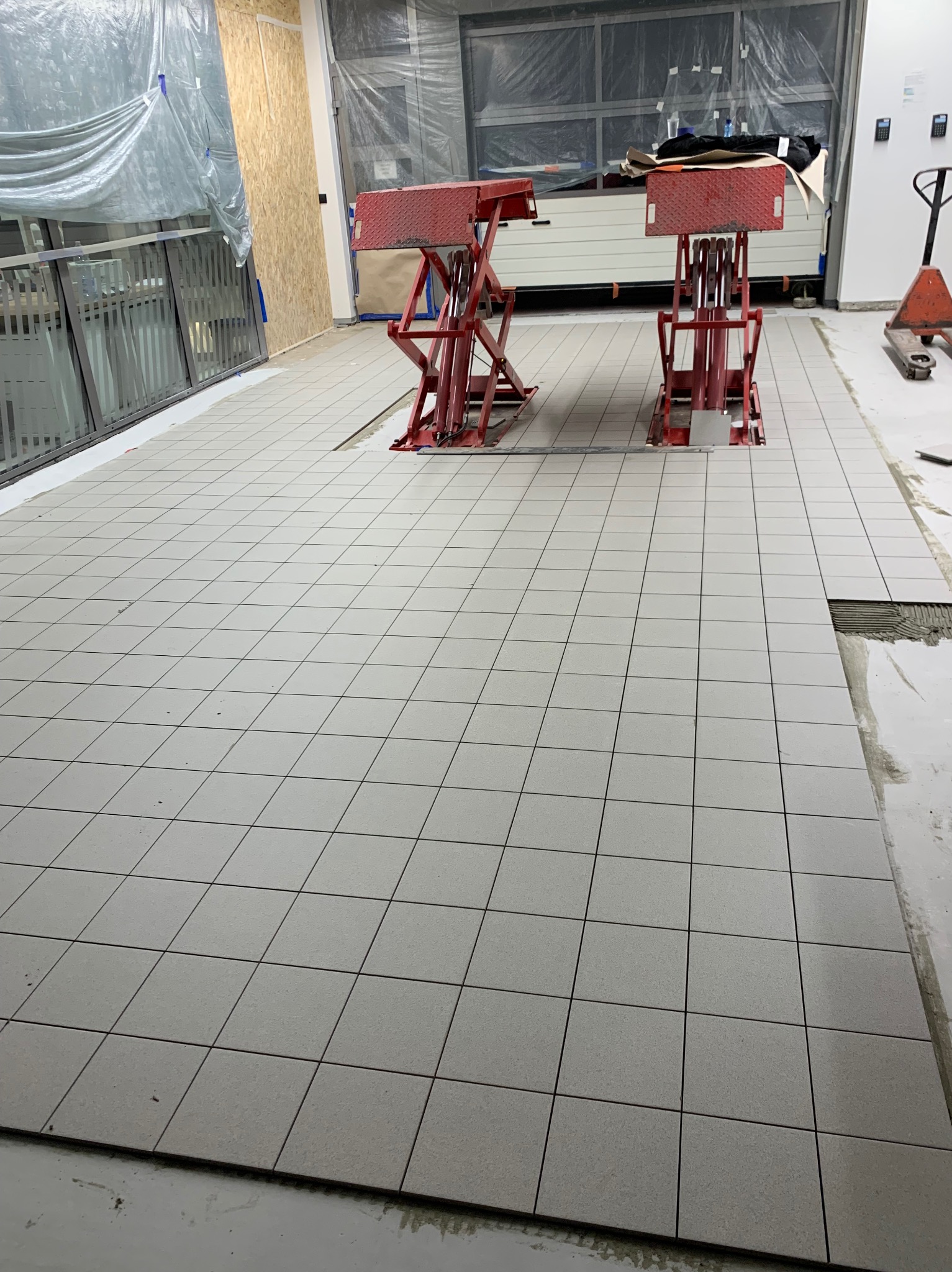 Układanie jasnoszarych, kwadratowych płytek podłogowych w garażu, z dwiema czerwonymi podnośnikami samochodowymi w tle, zabezpieczone folią ściany i brama garażowa.