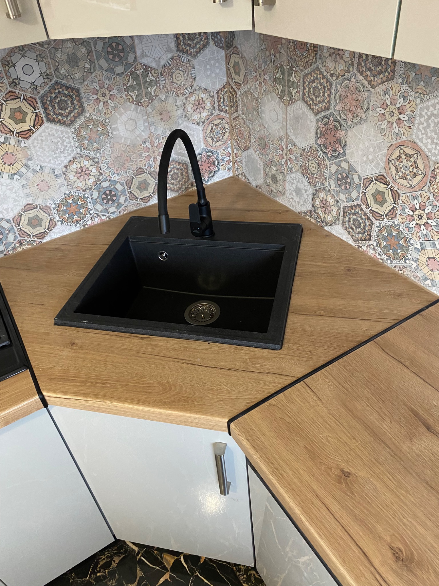 Czarny zlew z baterią na blacie imitującym drewno, nad którym widoczne są heksagonalne płytki w stylu patchwork. Uwagę przykuwa nietypowe połączenie wzorów i materiałów.