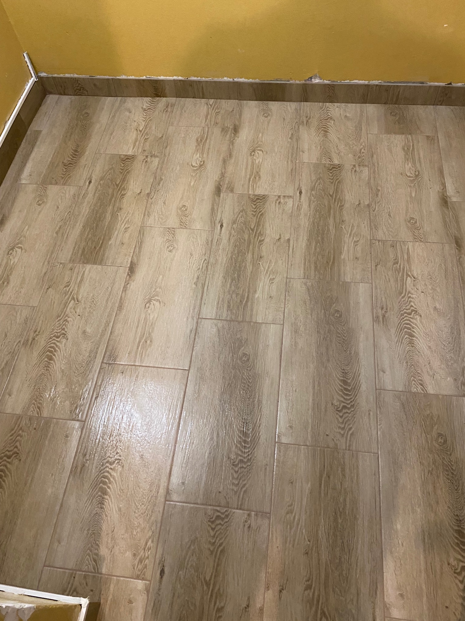 Ułożone, prostokątne płytki podłogowe imitujące drewno, z widocznymi fugami i fragmentem żółtej ściany w tle. Widoczny fragment listwy przypodłogowej.
