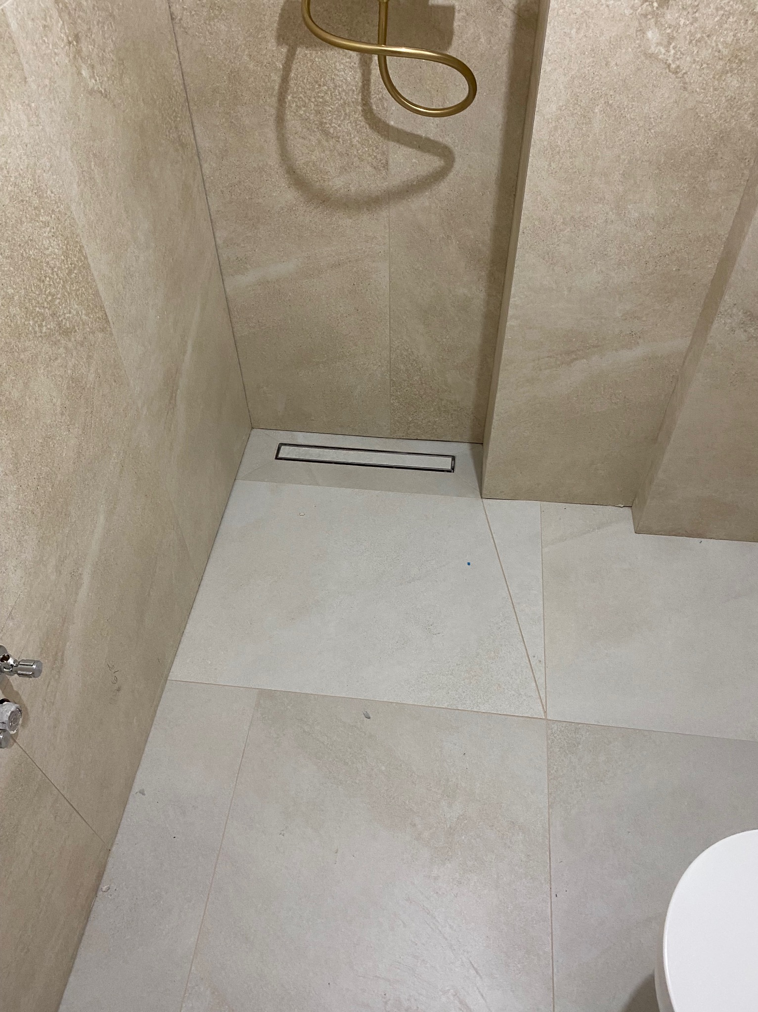 Beżowa łazienka z odpływem liniowym i złotą słuchawką prysznicową. Płytki imitujące kamień na ścianach i podłodze. Minimalistyczny design.