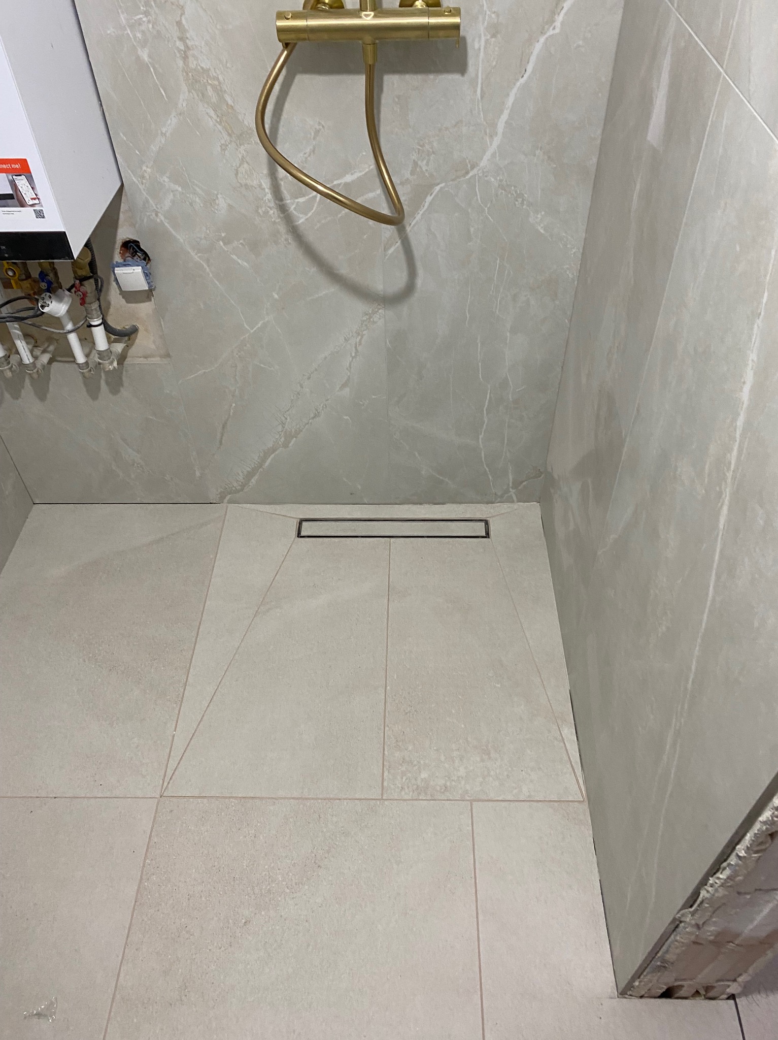 Jasne, marmurowe płytki w kabinie prysznicowej z odpływem liniowym. Widoczne złote elementy armatury i fragment bojlera. Minimalistyczny design.