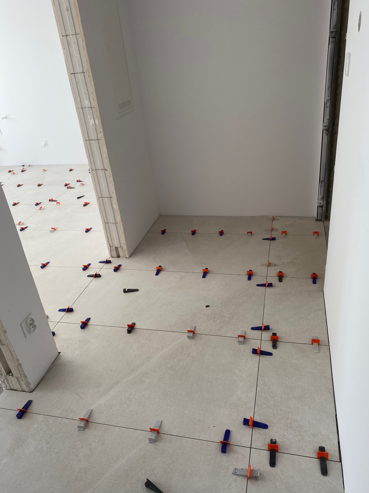 Jasne płytki podłogowe w trakcie układania, z użyciem klipsów poziomujących i sznurka, widok z góry, surowe wnętrze w trakcie remontu.