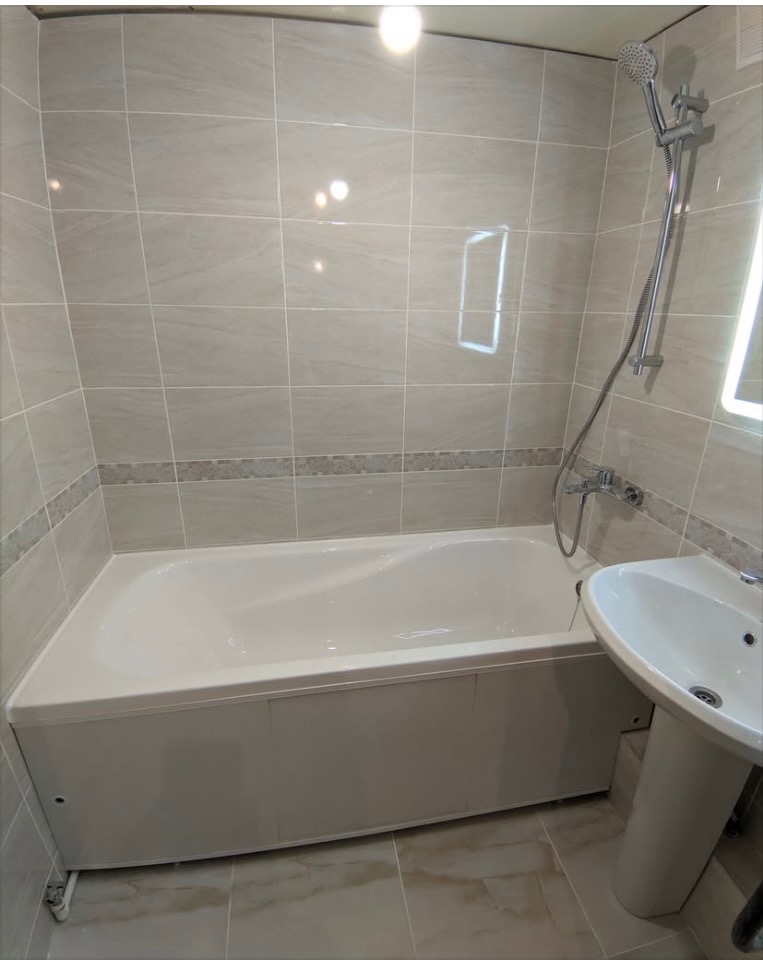 Jasna łazienka po remoncie: beżowe płytki na ścianach i podłodze, biała wanna z obudową, umywalka na postumencie, chromowana armatura. Minimalistyczny design.