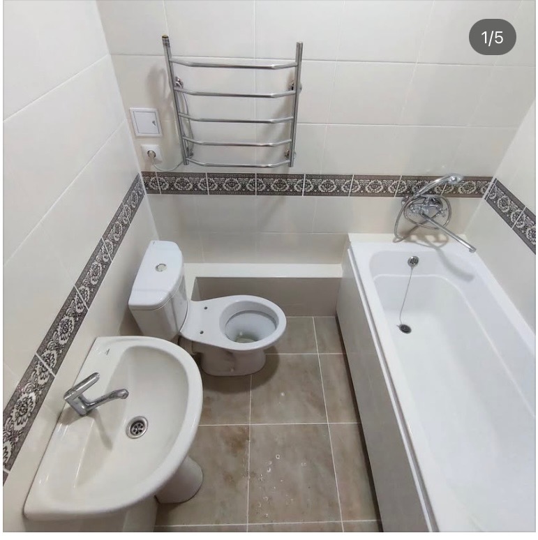 Ukończona łazienka z beżowymi płytkami, białą armaturą (umywalka, toaleta, wanna) i grzejnikiem drabinkowym. Dekoracyjny pas płytek na ścianach.