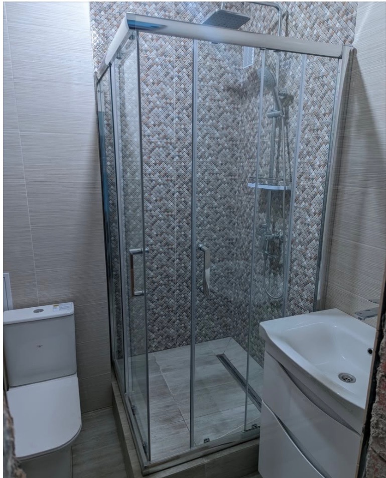 Wykończona łazienka z kabiną prysznicową z chromowanymi profilami, umywalką z szafką i toaletą. Na ścianach mozaika i płytki imitujące drewno.