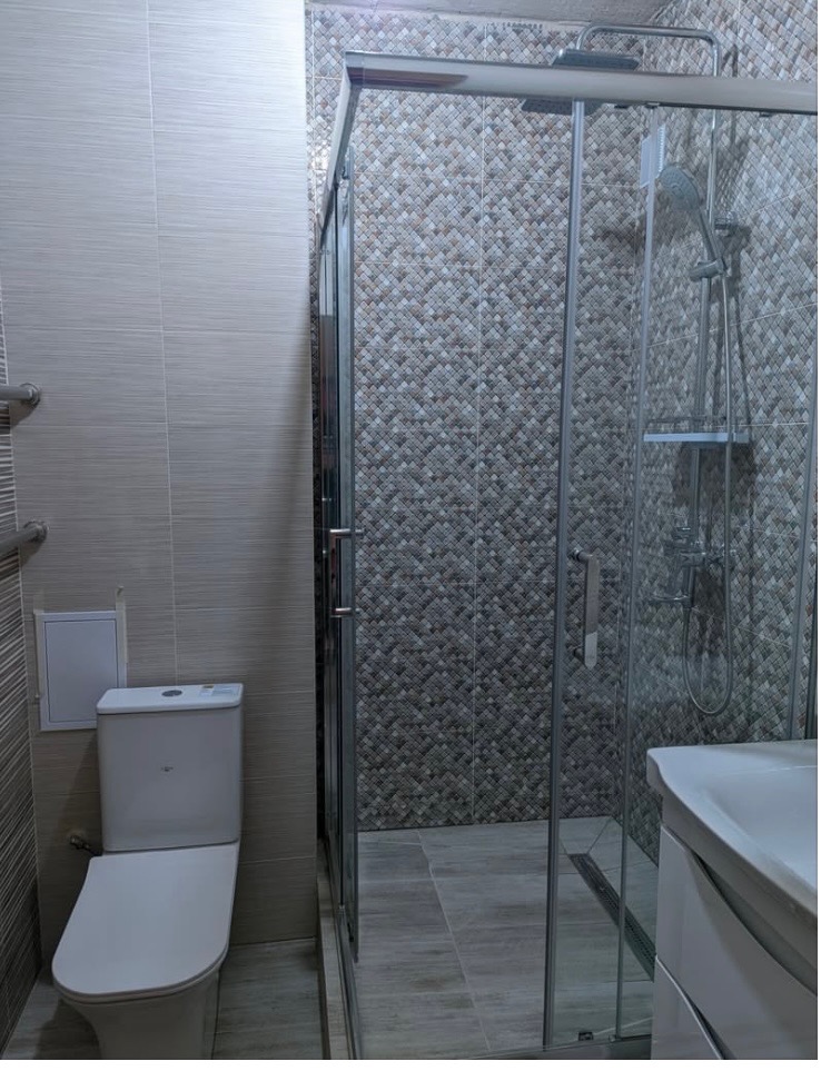 Wykończona łazienka z kabiną prysznicową, toaletą i umywalką. Ściany wyłożone płytkami w różnych odcieniach szarości i beżu. Nowoczesny design.