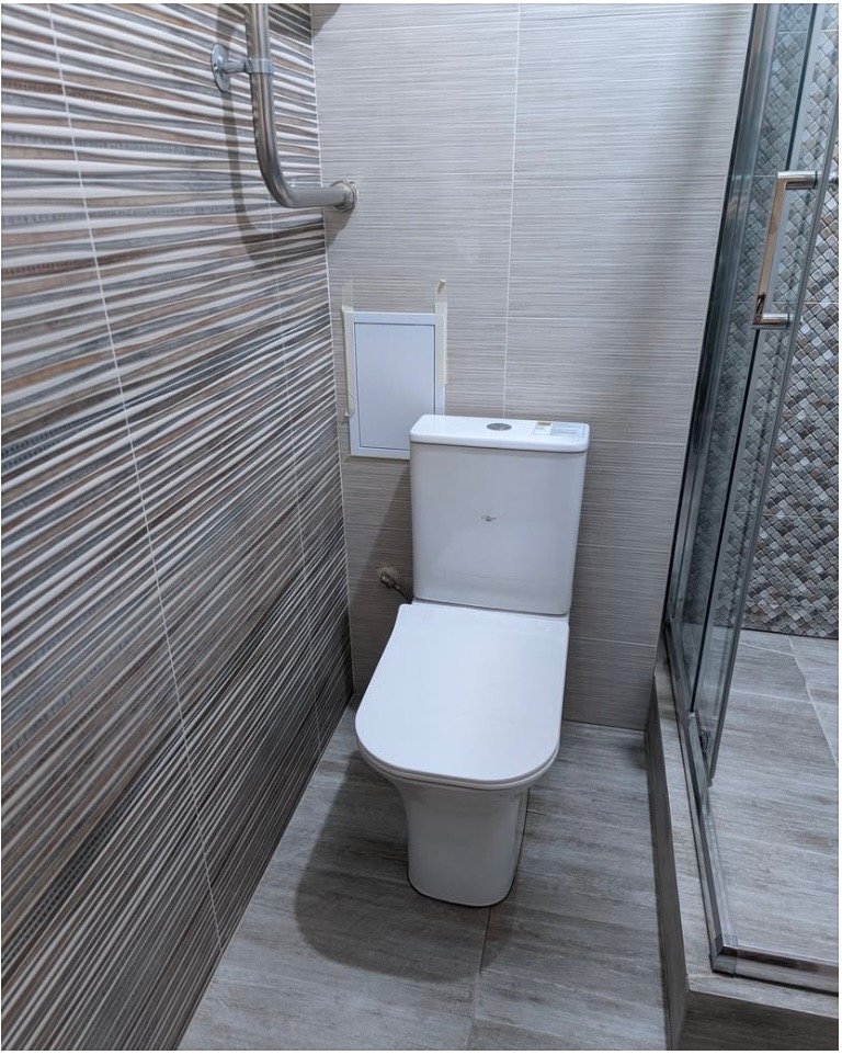 Wykończona łazienka z toaletą, szarobrązowe płytki na ścianach i podłodze, kabina prysznicowa z widoczną mozaiką wewnątrz, rura i rewizja na ścianie.