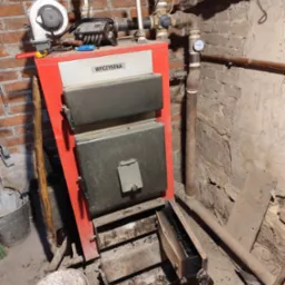 Czerwony piec centralnego ogrzewania w kotłowni, z otwartymi drzwiczkami paleniska, widoczne rury instalacji, narzędzia i pojemnik z popiołem, surowe ściany z cegły i betonu.