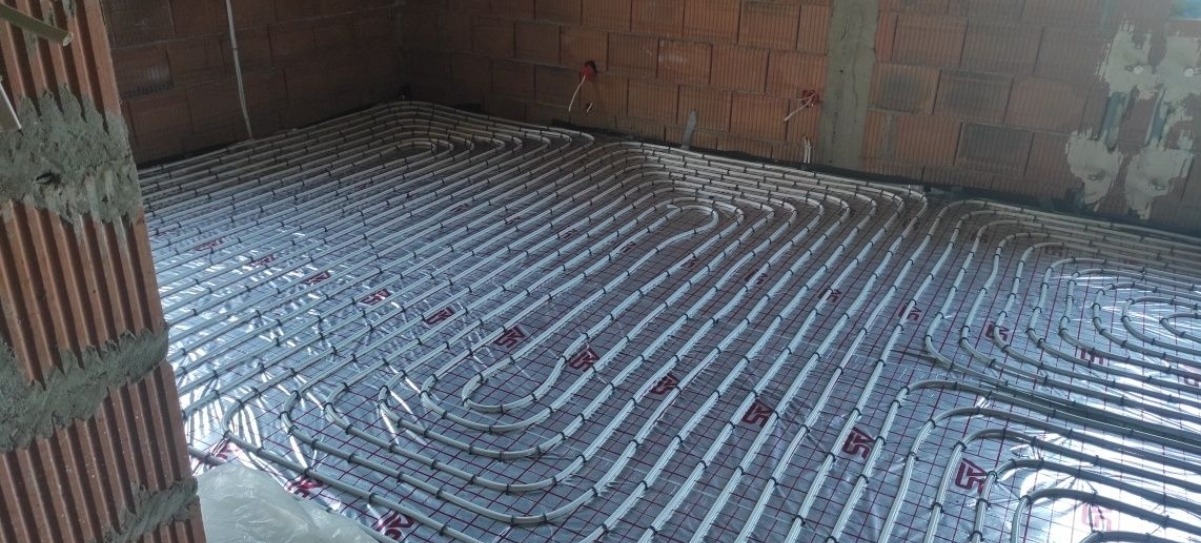 Instalacja ogrzewania podłogowego z rur PEX ułożonych na folii izolacyjnej z nadrukowaną siatką, widok na surową ceglaną ścianę w tle.