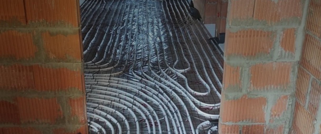 Układ rur ogrzewania podłogowego na folii izolacyjnej, widoczne z perspektywy otworu w surowej ścianie z cegły dziurawki.