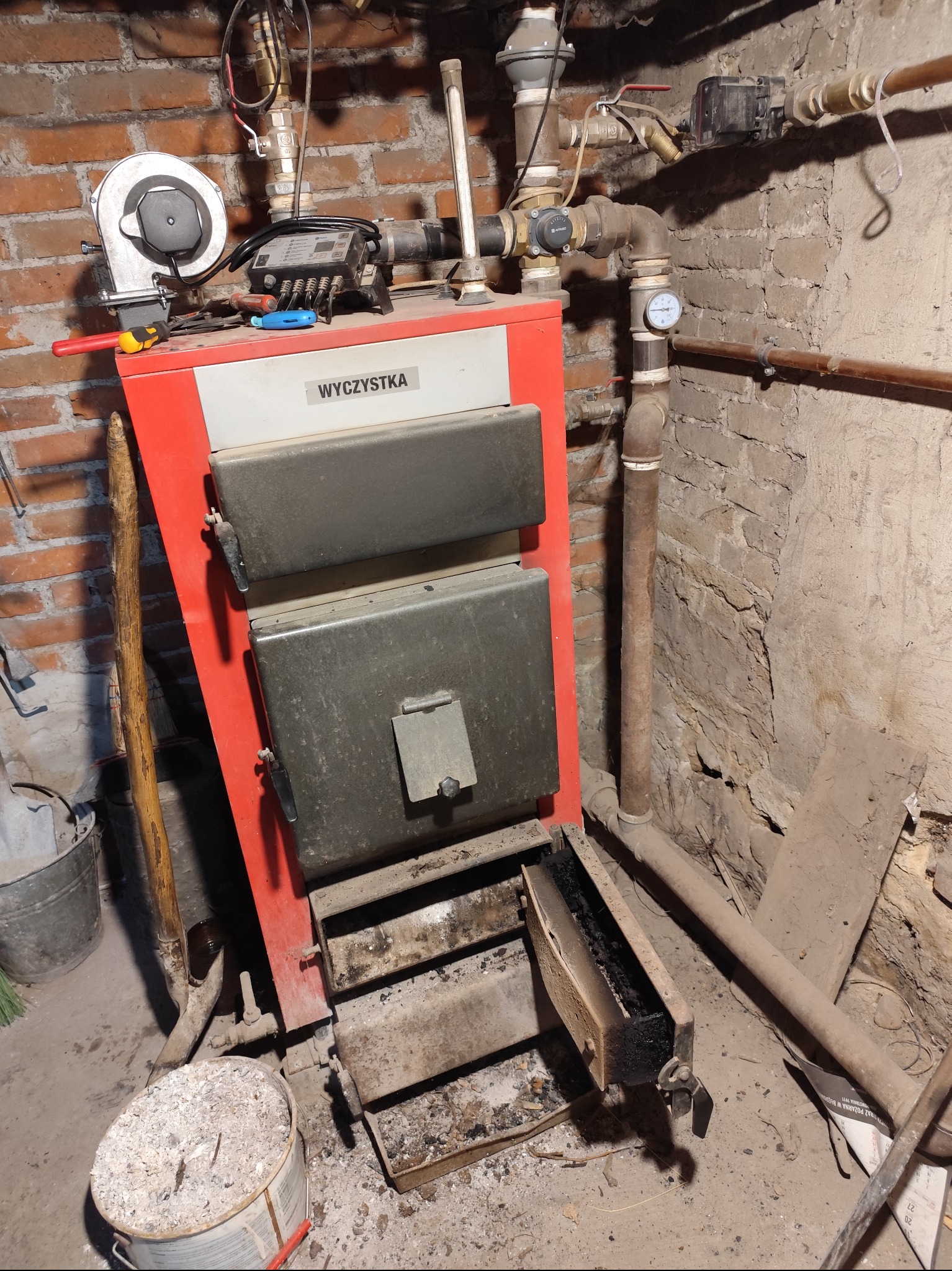 Czerwony piec centralnego ogrzewania w kotłowni, z otwartymi drzwiczkami paleniska, widoczne rury instalacji, narzędzia i pojemnik z popiołem, surowe ściany z cegły i betonu.