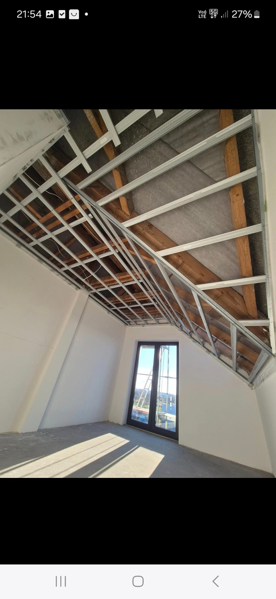 Skośne poddasze z oknem, w trakcie montażu metalowej konstrukcji sufitu podwieszanego. Widoczne drewniane elementy więźby dachowej i surowe ściany.