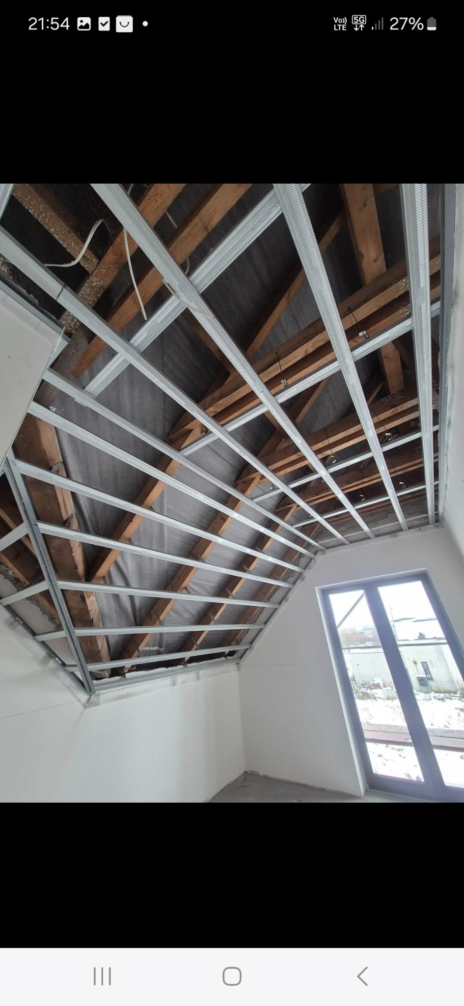 Szkielet sufitu podwieszanego z metalowych profili, widoczne drewniane elementy konstrukcyjne dachu, okno w skosie dachu, białe ściany. Etap prac wykończeniowych.